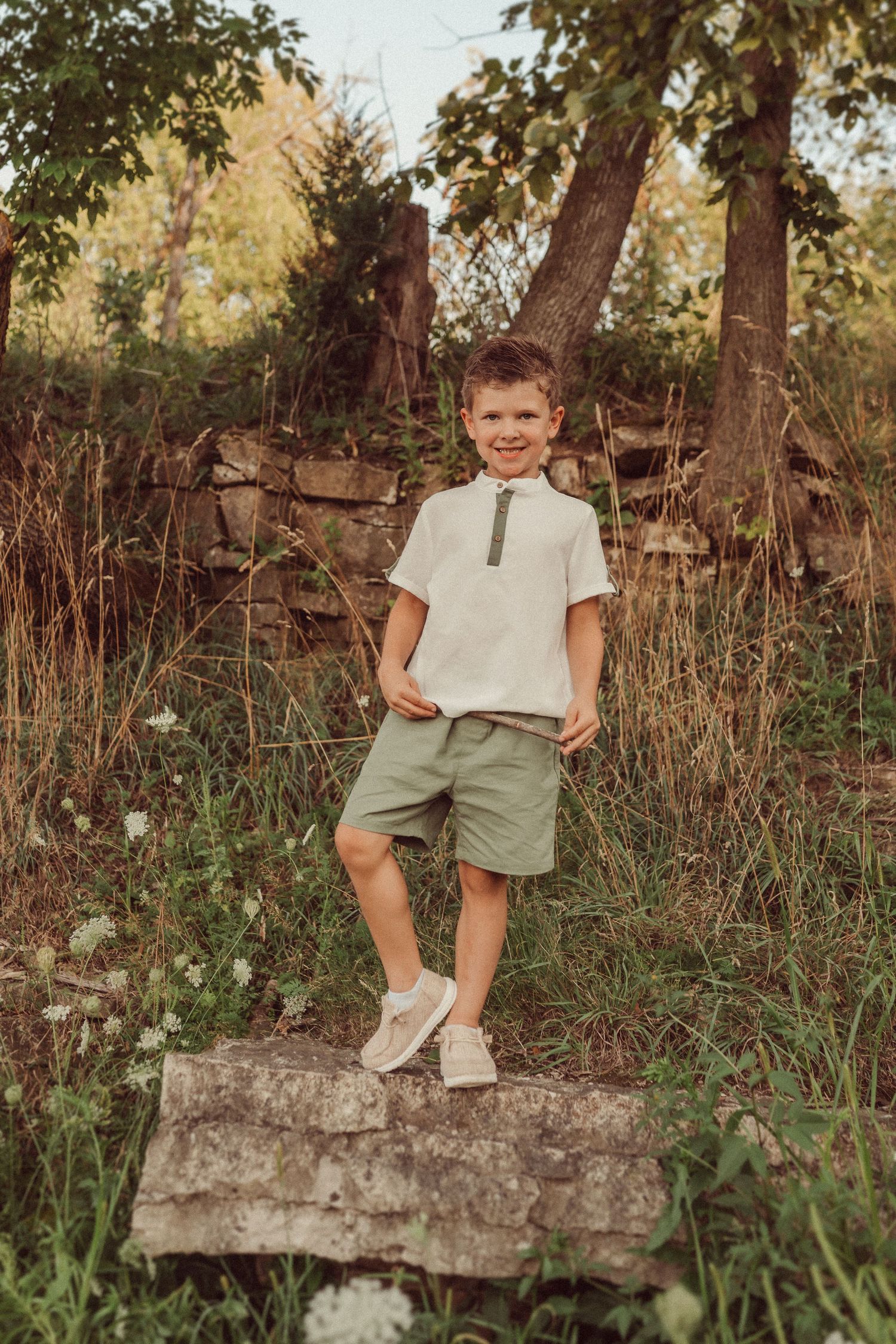 Child stands confidently on stone ledge wearing casual summer outfit in natural woodland setting.