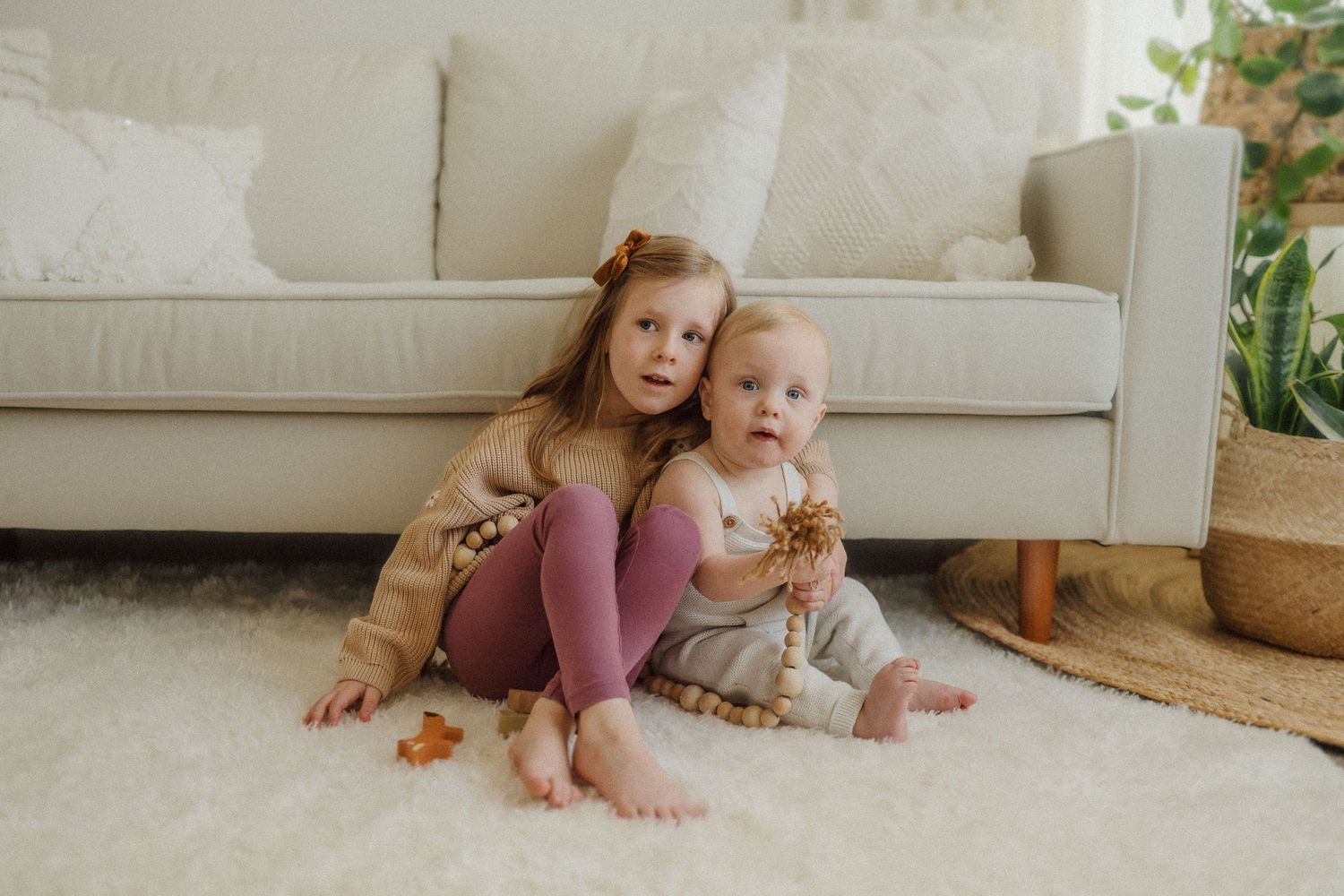 Two young children sit together on a white carpet in front of a light-colored couch.