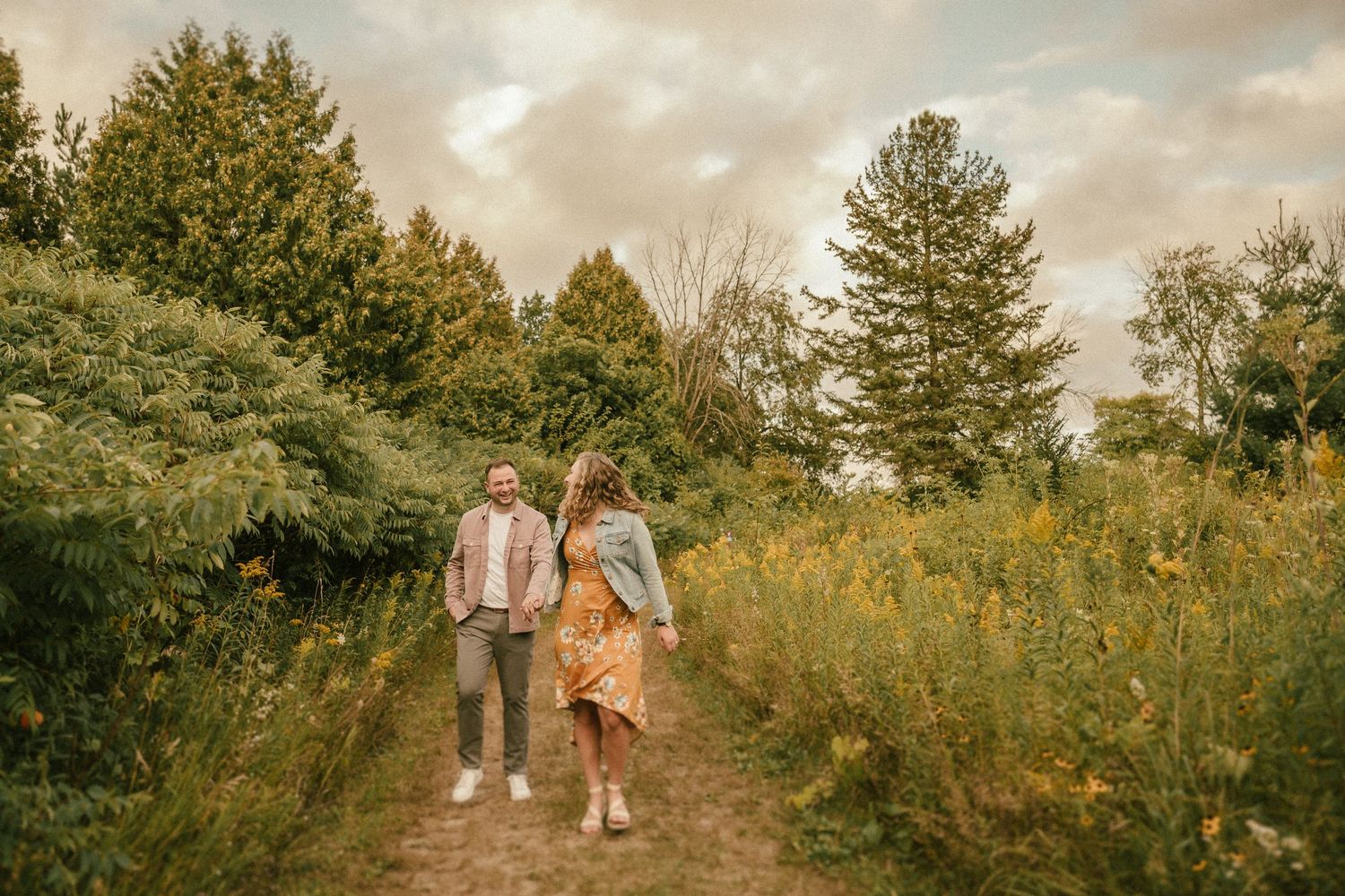 A couple walks hand-in-hand down a wilderness trail surrounded by evergreen trees and wildflowers.