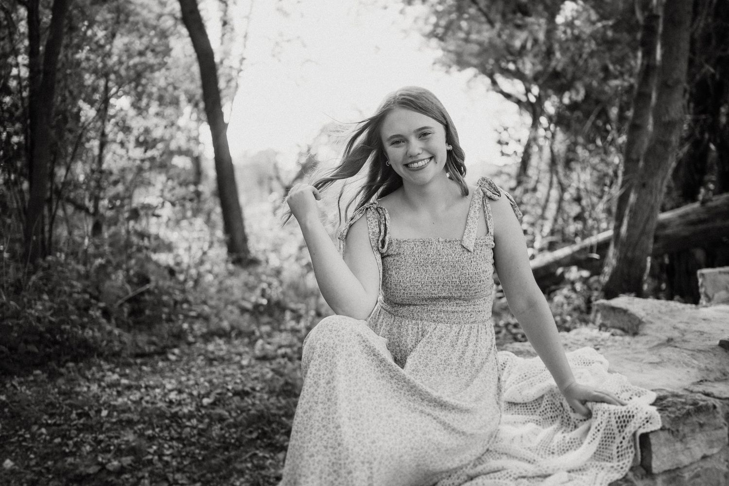 Black and white photo of a woman in flowing dress twirling outdoors by stone wall.