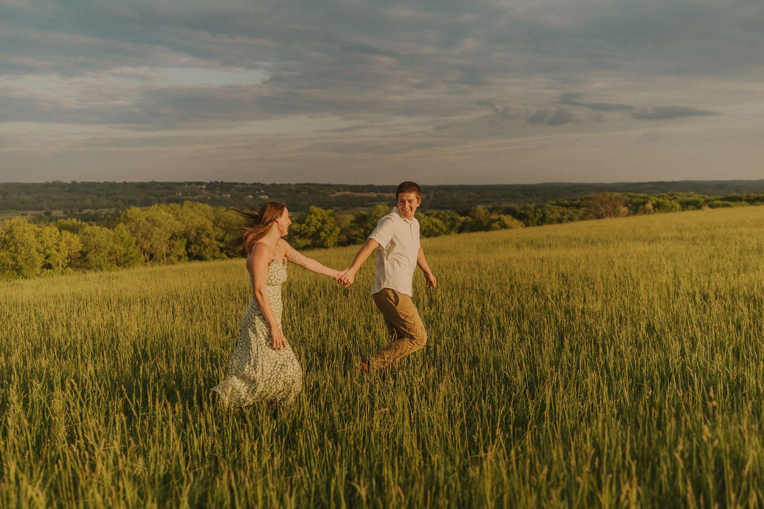 A couple runs playfully through a golden wheat field at sunset with rolling hills in the background.
