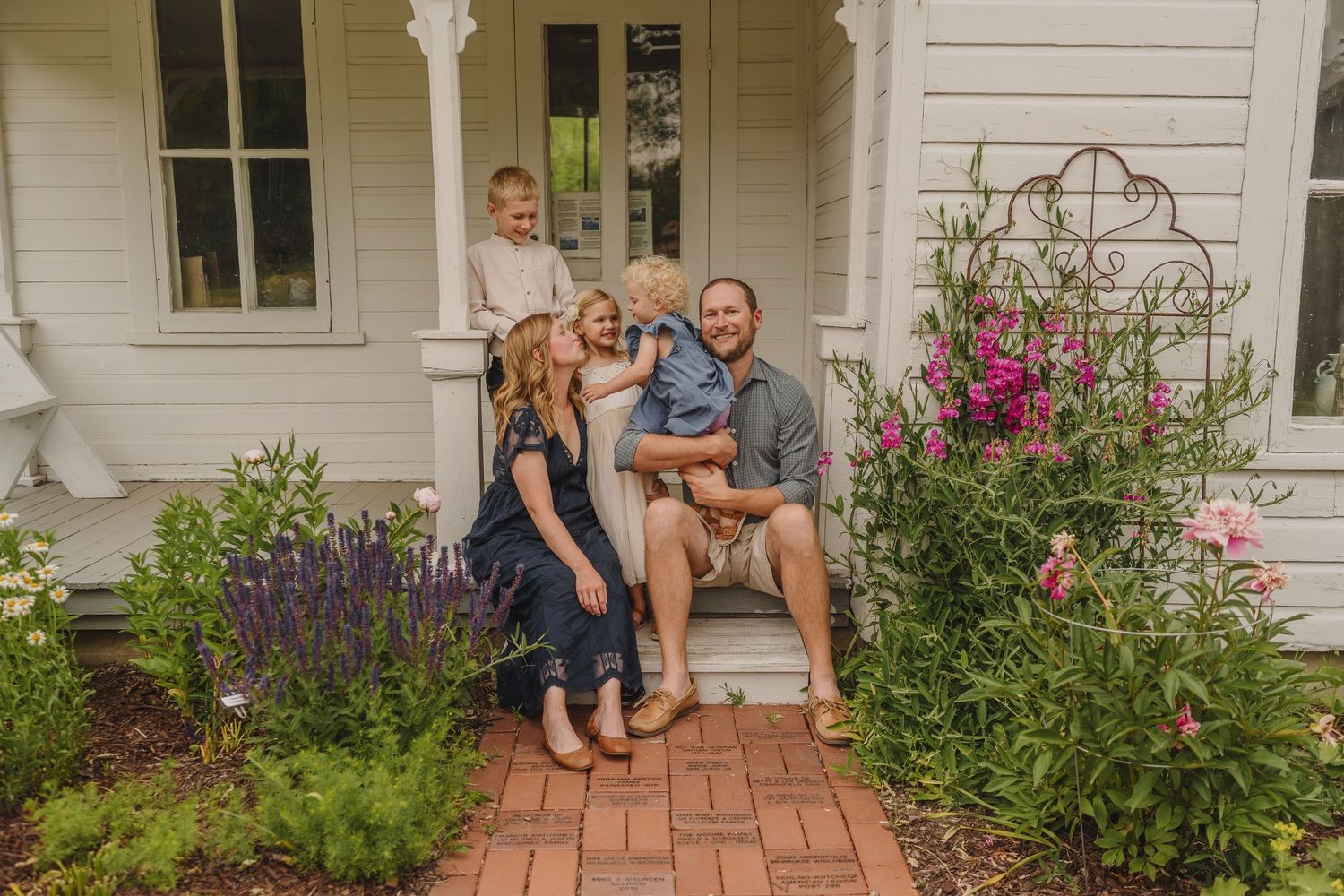 A group gathers on brick steps of a white farmhouse porch surrounded by vibrant purple and pink flowers in bloom.