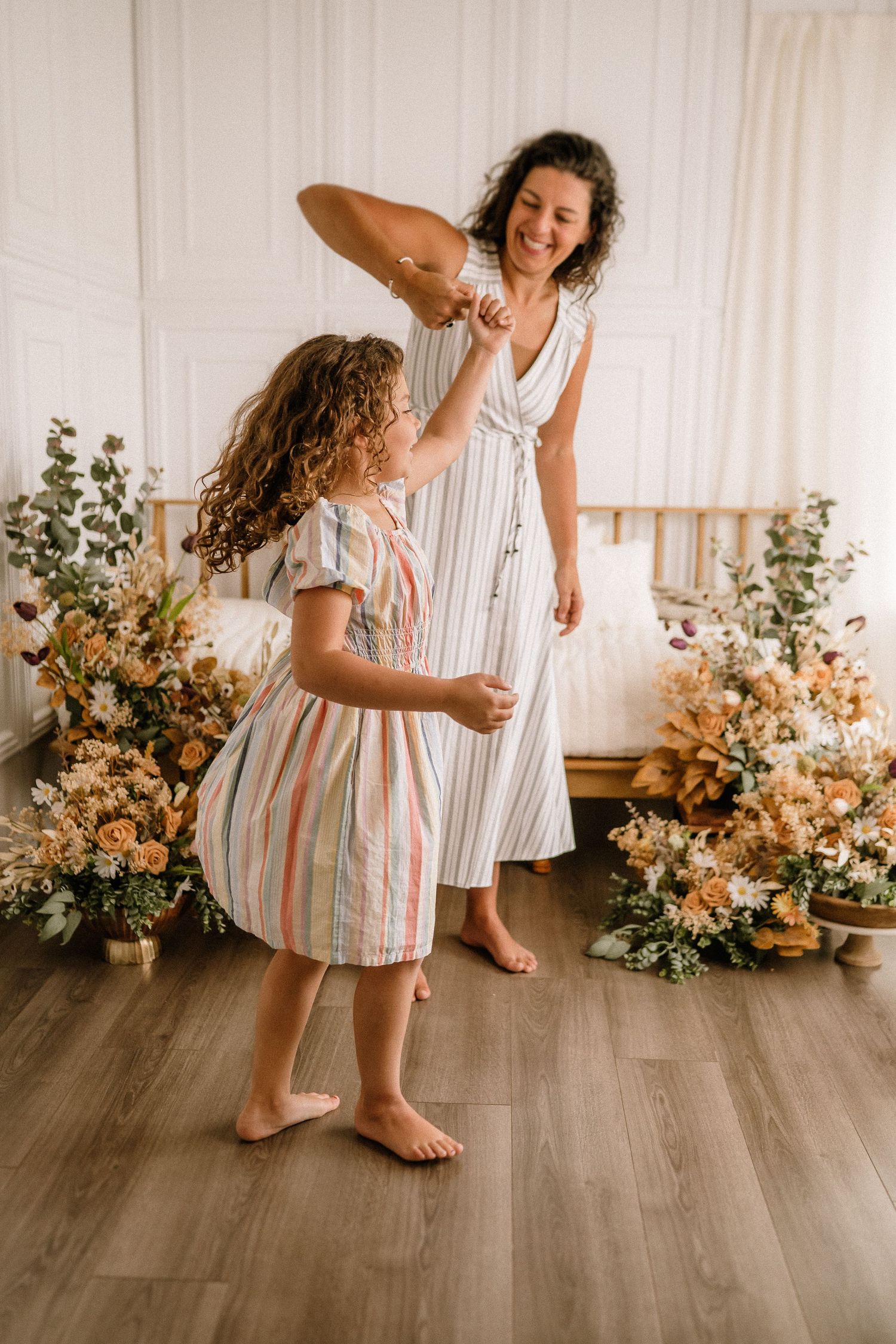 Two people in white and striped summer dresses dance barefoot together in a room decorated with floral arrangements.