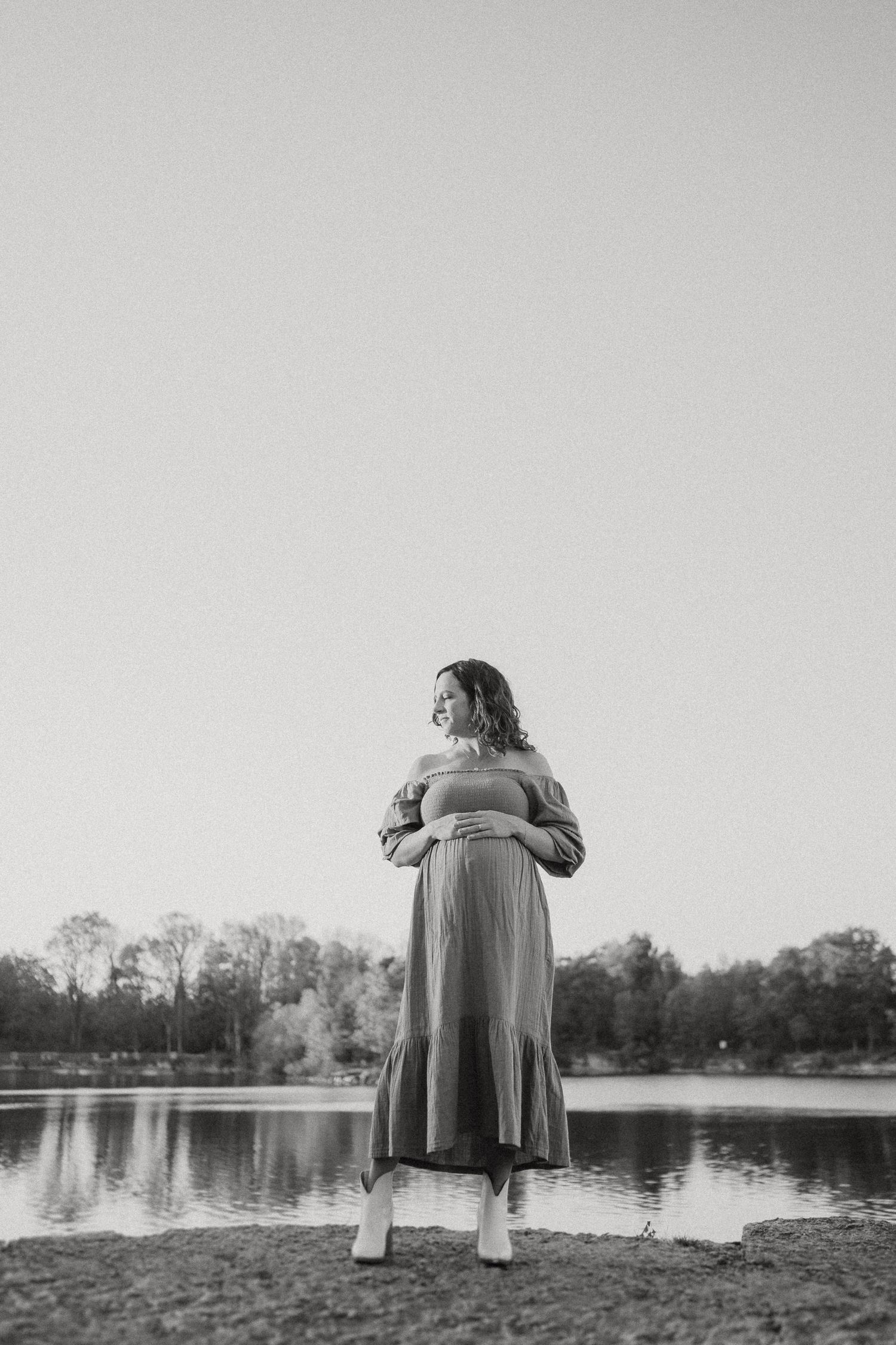Black and white photo of a pregnant woman in a maxi dress standing by a lake.