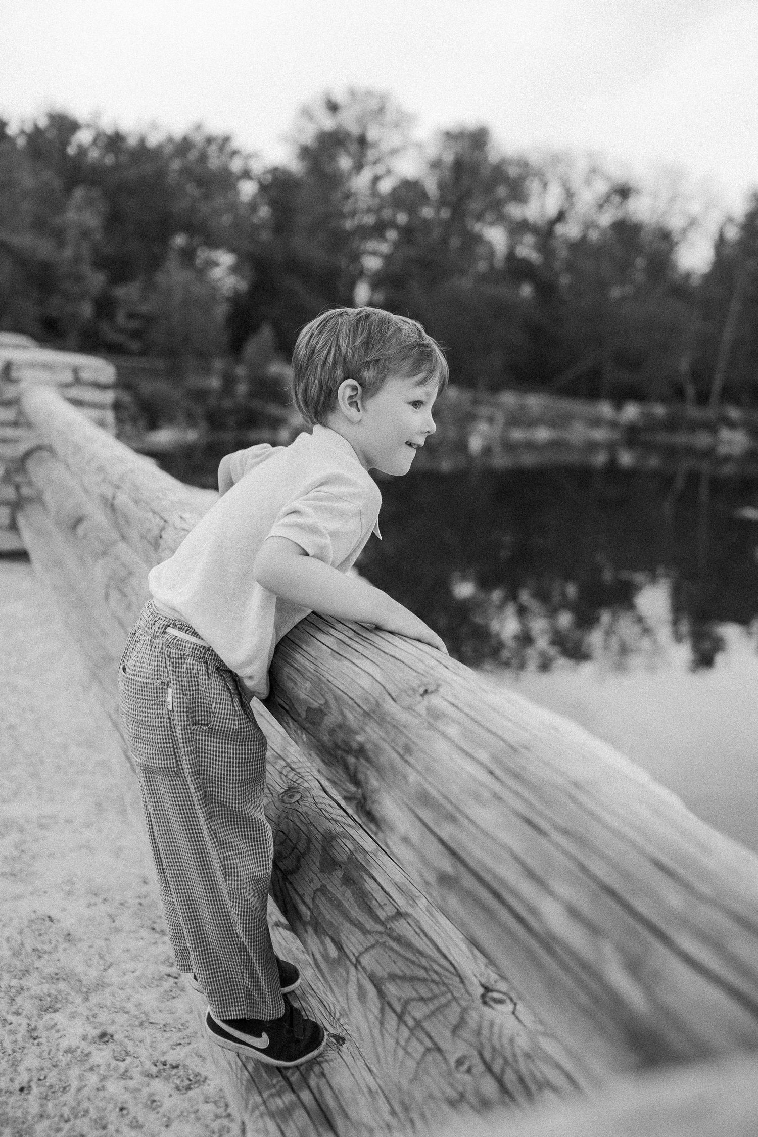 A black and white photo of a child leaning over a wooden railing near a lake with trees in the background.