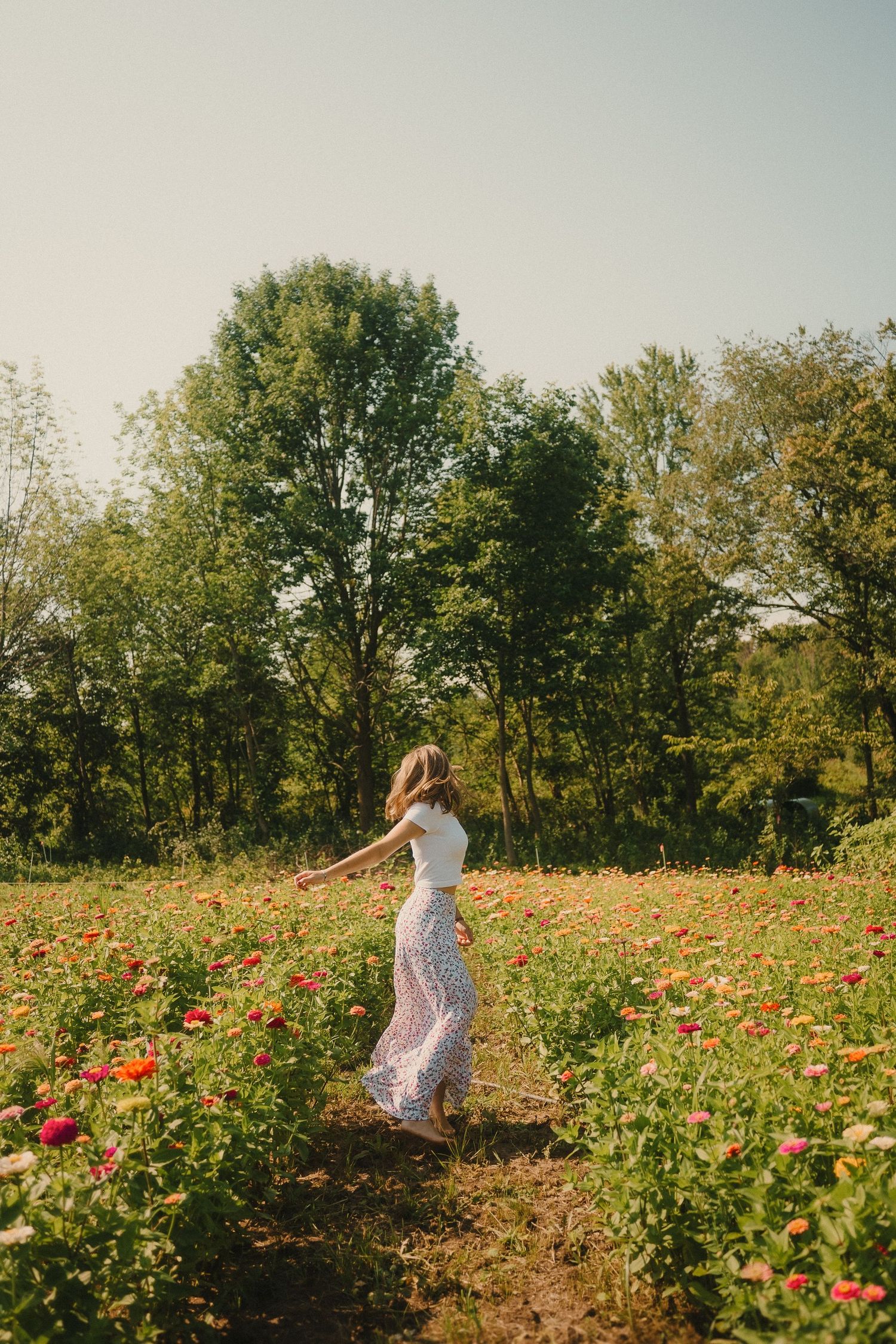 A woman in a white dress dances joyfully through a blooming wildflower field on a sunny summer day.