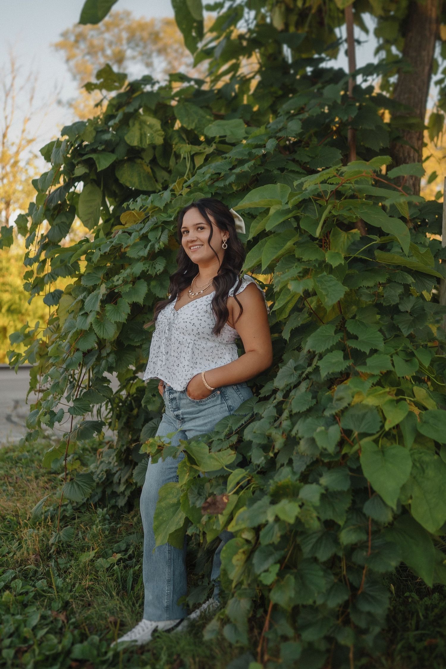 A person poses outdoors by lush green foliage wearing a floral top and jeans on a sunny summer evening.