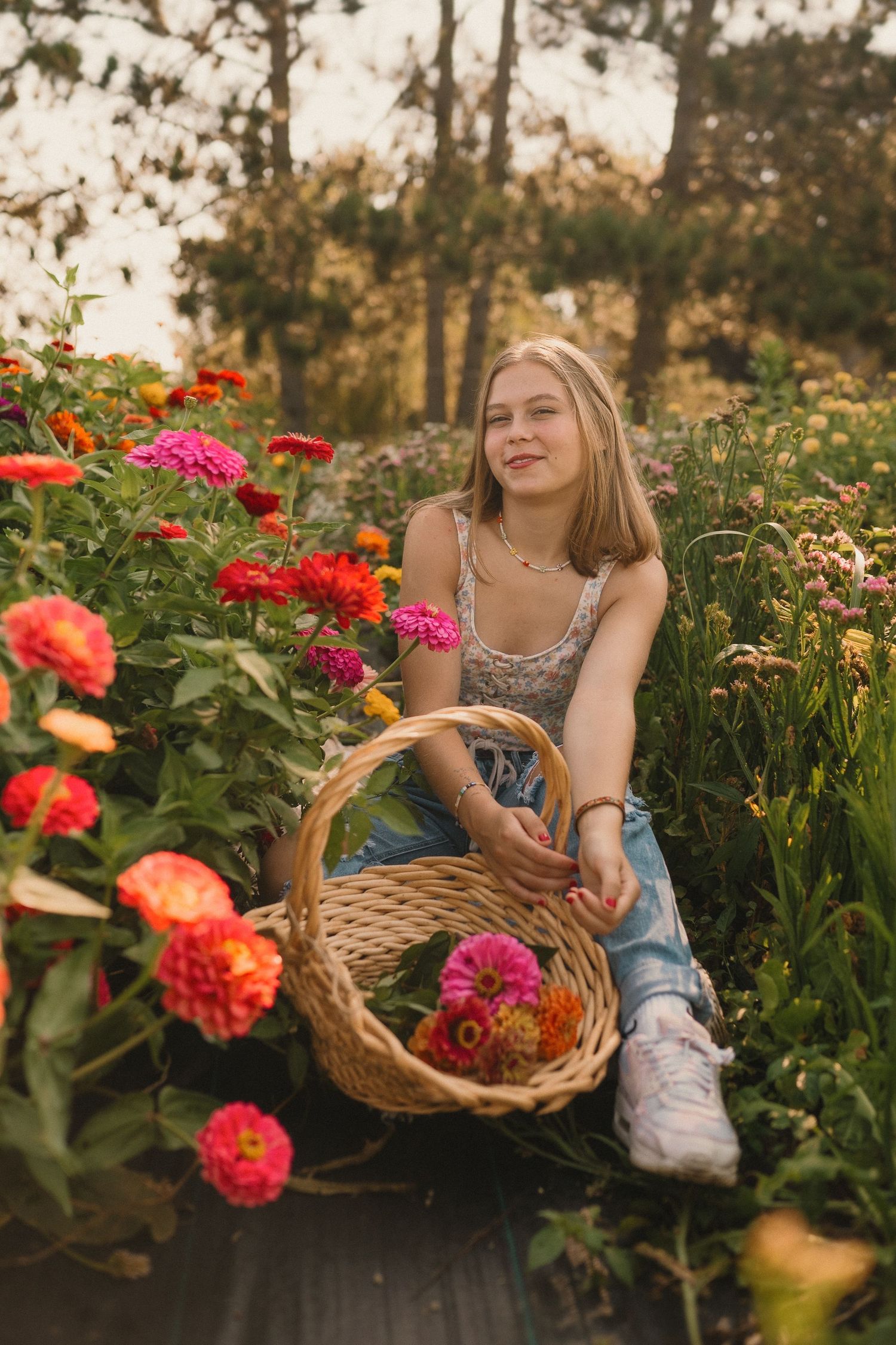 Someone holding wicker basket while sitting among colorful zinnias and dahlias in garden at dusk.