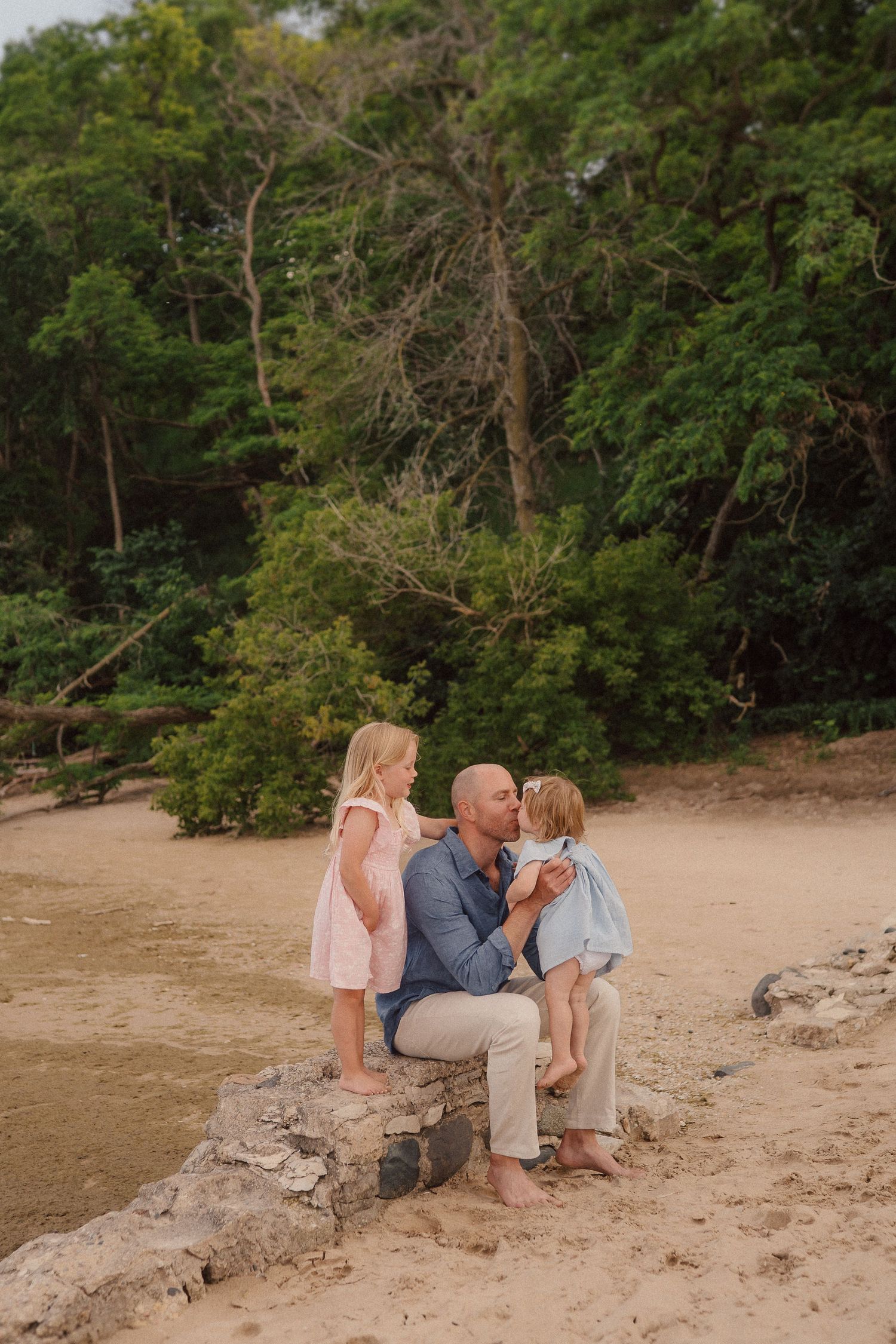 Family enjoying a playful moment together sitting on sandy rocks at a wooded beach during sunset.