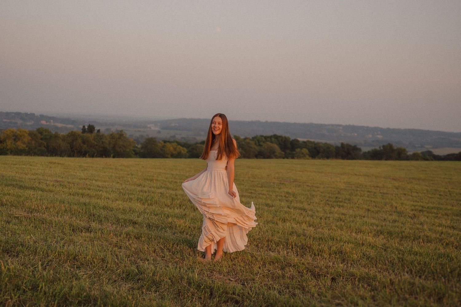 A person in a flowing peach dress walks through a golden wheat field at sunset in the countryside.