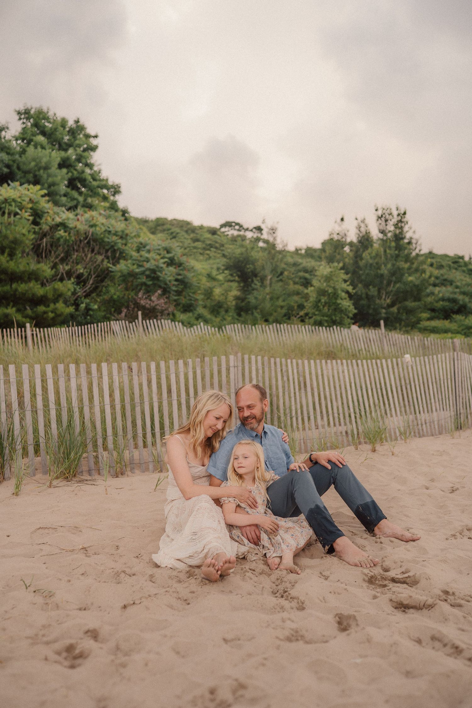 A casual family portrait on a beach with wooden fence and sand dunes on an overcast summer day.