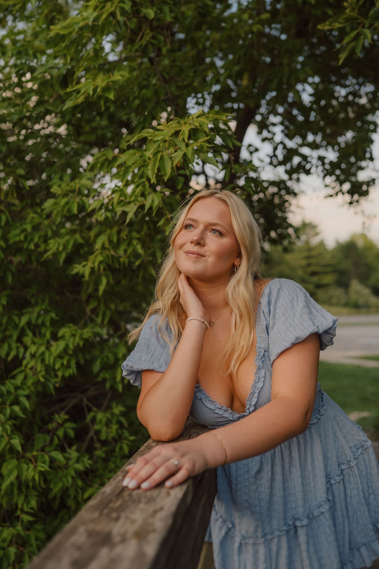 Person in blue dress poses thoughtfully against lush green foliage.
