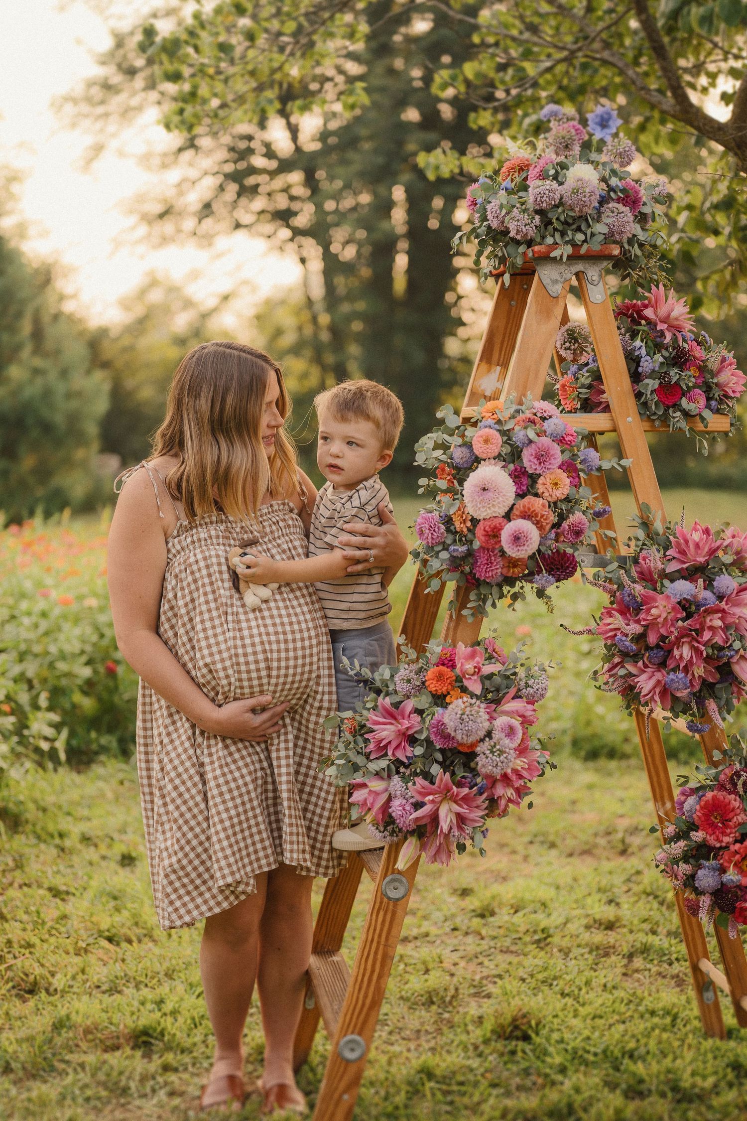A pregnant woman in a checkered dress holds a child beside a wooden ladder decorated with pink and purple floral arrangements.