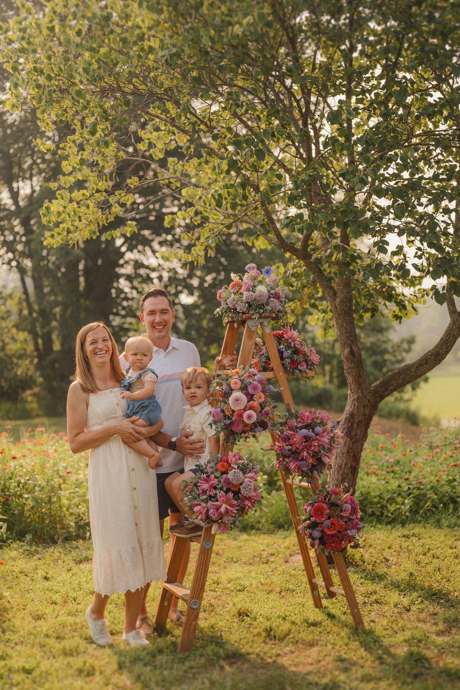 A family poses near a floral-decorated wooden ladder in a sunlit garden during golden hour.