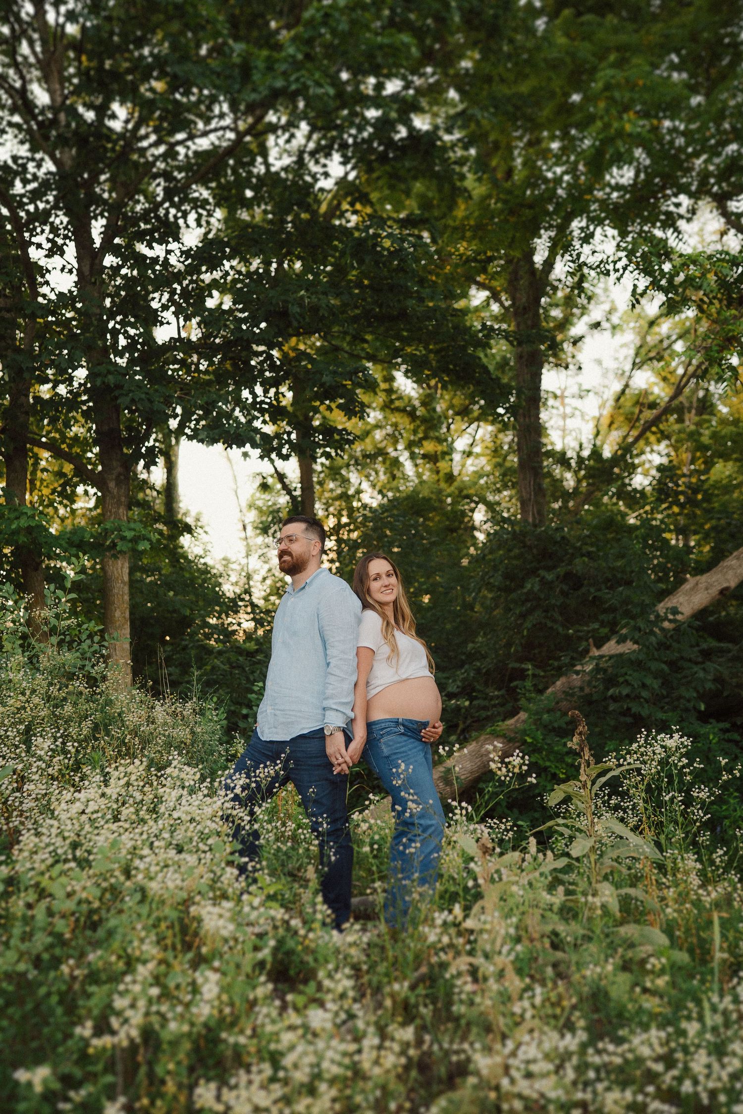 A couple walks together through a wildflower meadow surrounded by green trees at golden hour.
