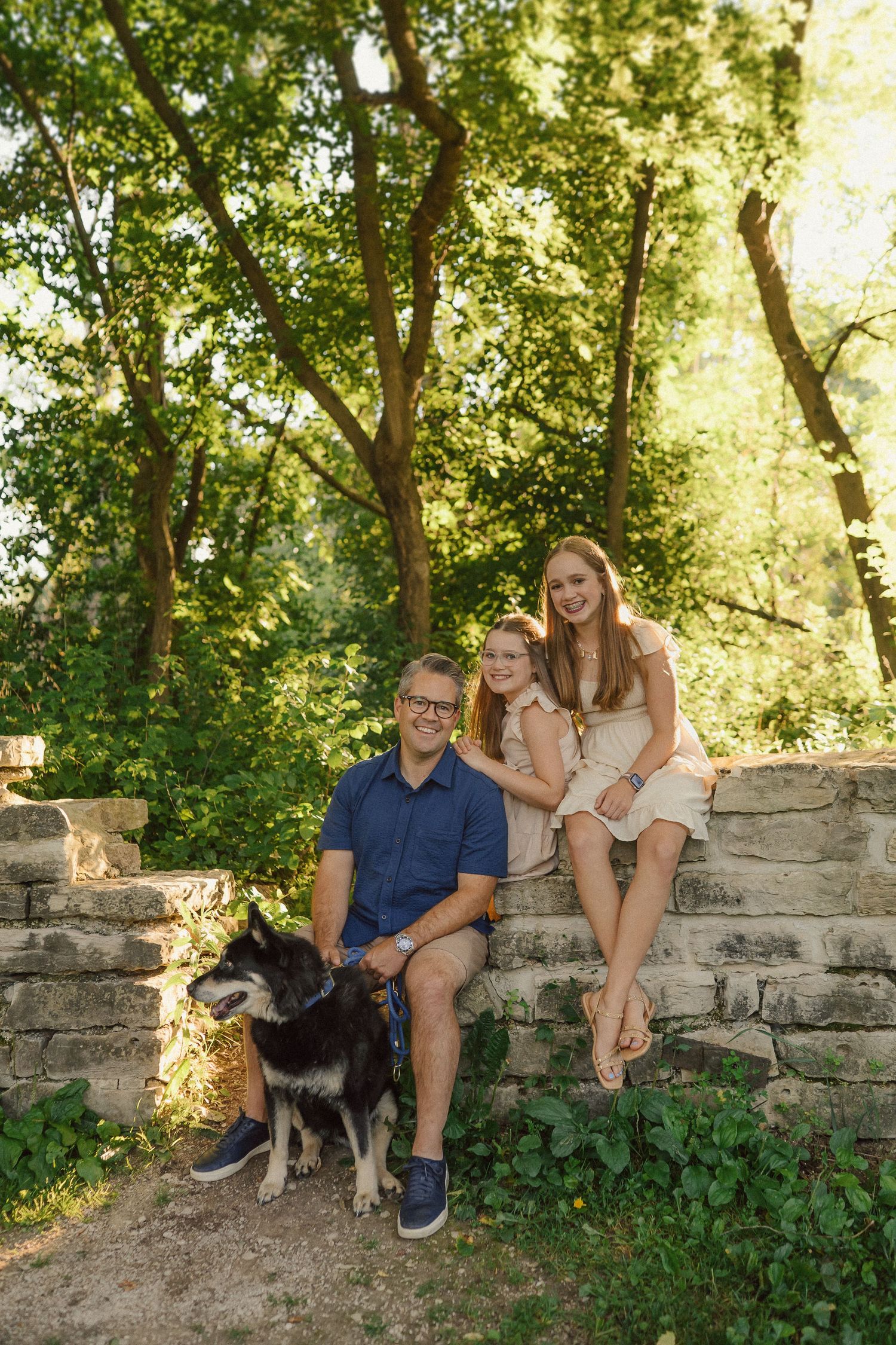 A family relaxes on stone steps surrounded by lush greenery with their black dog beside them.