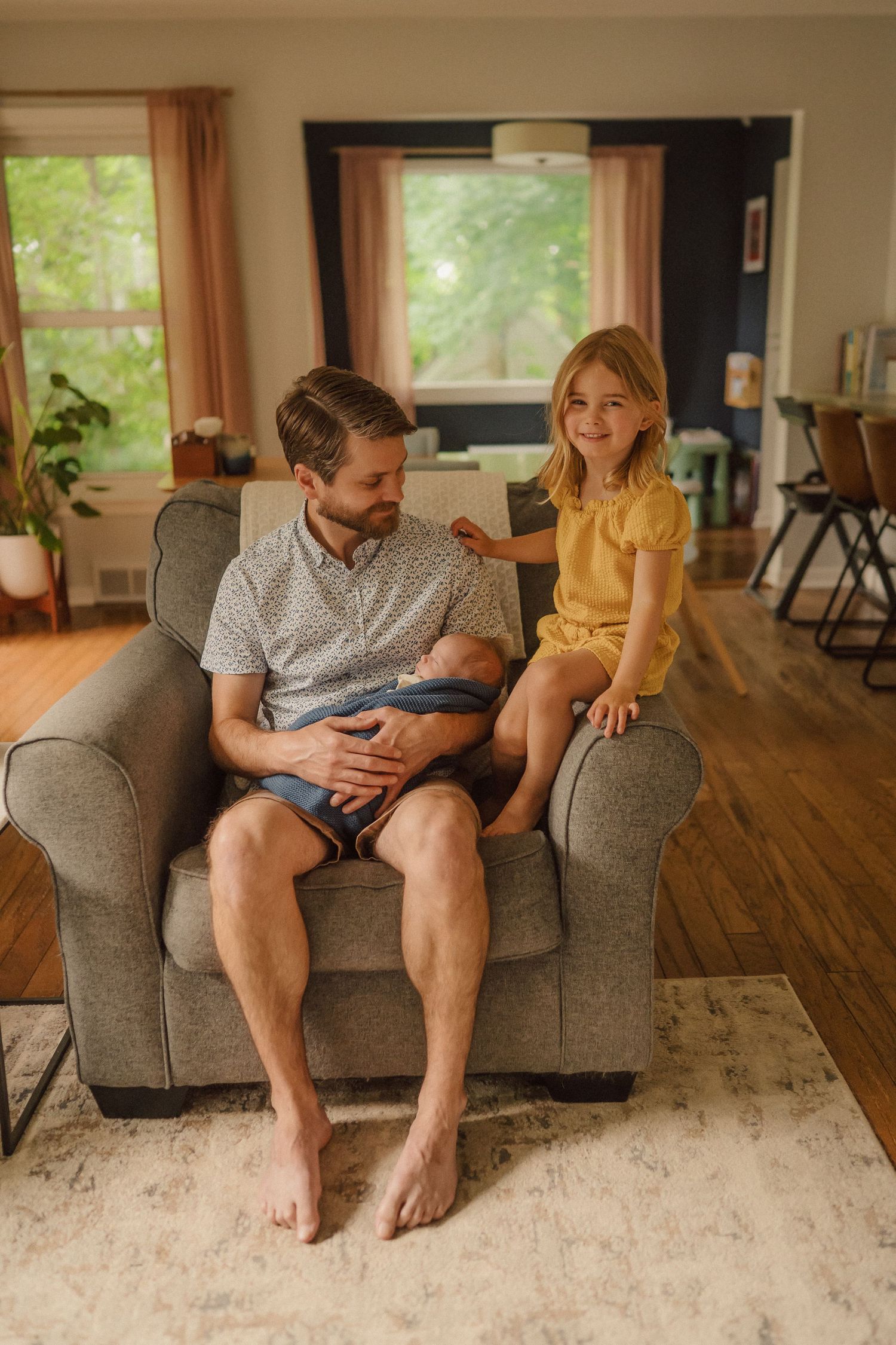 Intimate family moment in living room as adults sit on grey couch with newborn on sunny afternoon.