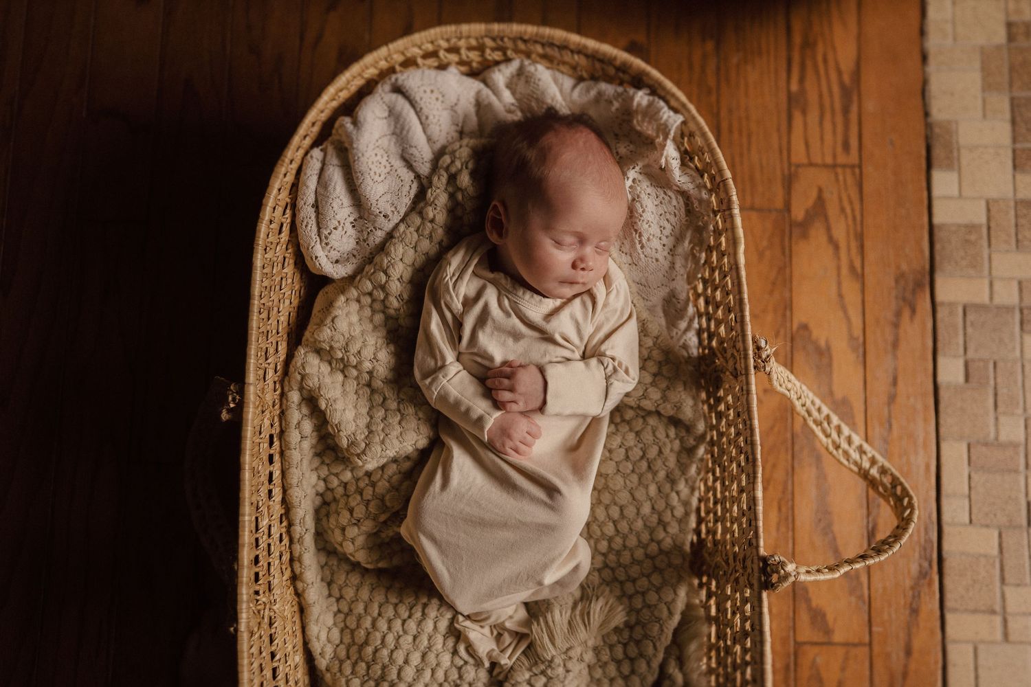 Sleeping newborn in beige swaddle blanket rests peacefully in woven bassinet on wooden floor.