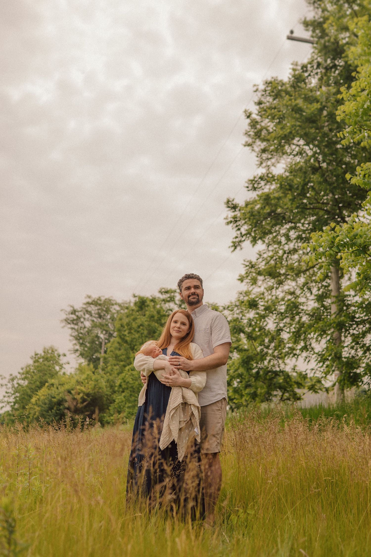 Family standing together in tall grass field with cloudy sky and tree backdrop.