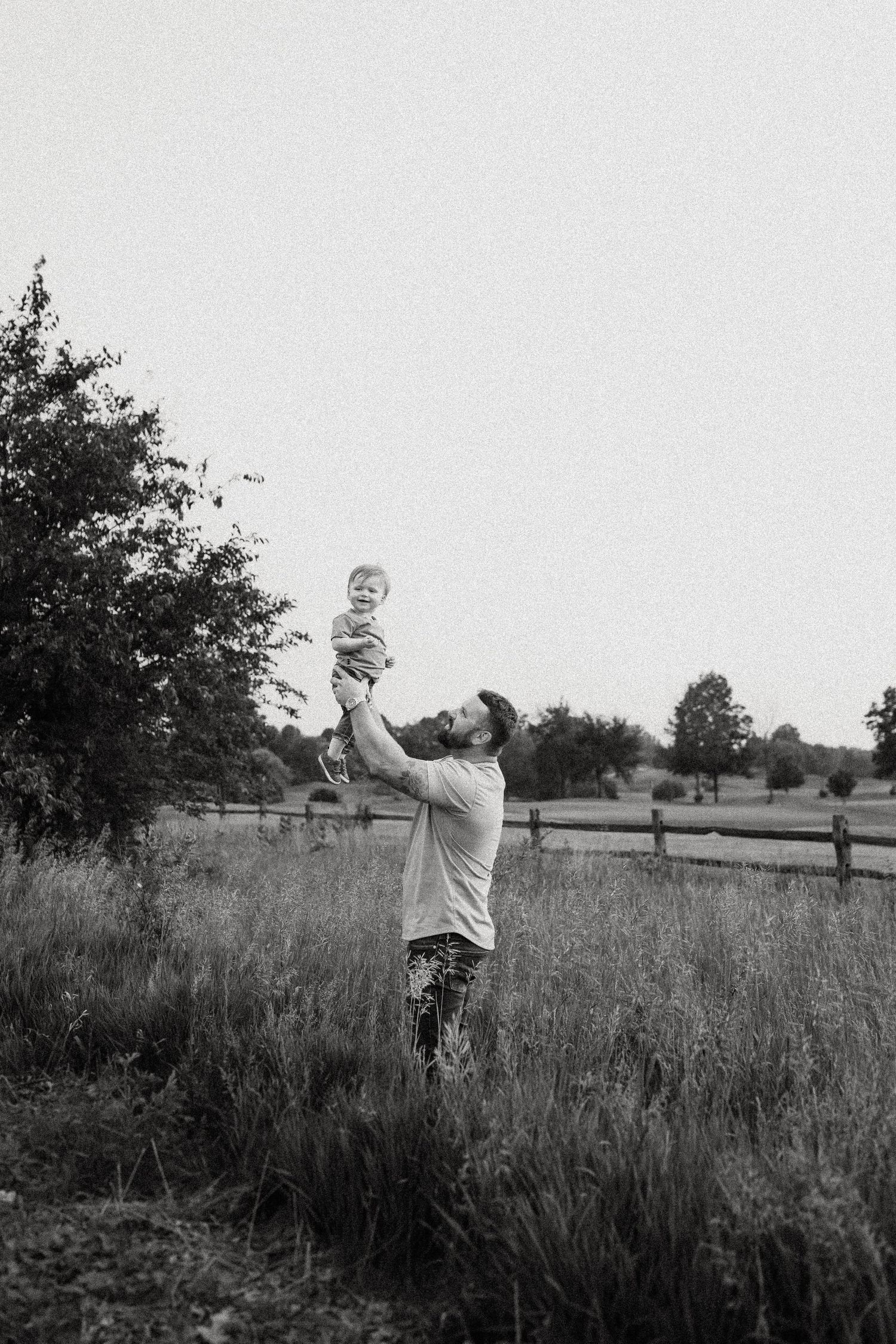 Black and white image capturing a playful moment between family members in a field with trees.