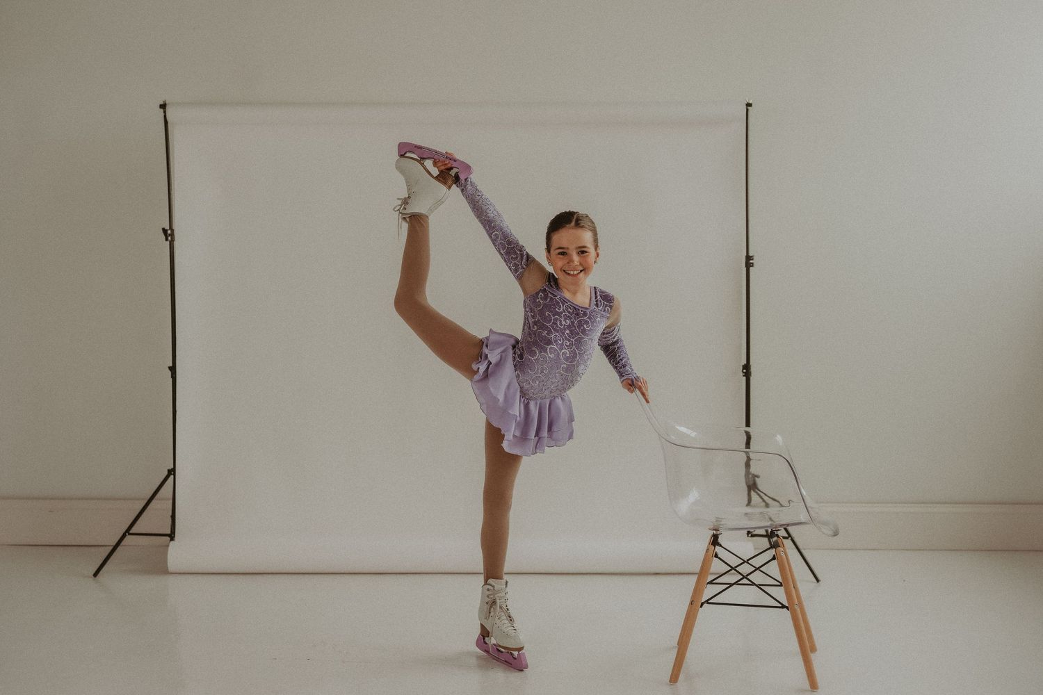 Figure skater in purple dress executes leg raise pose in front of white backdrop with modern chair.