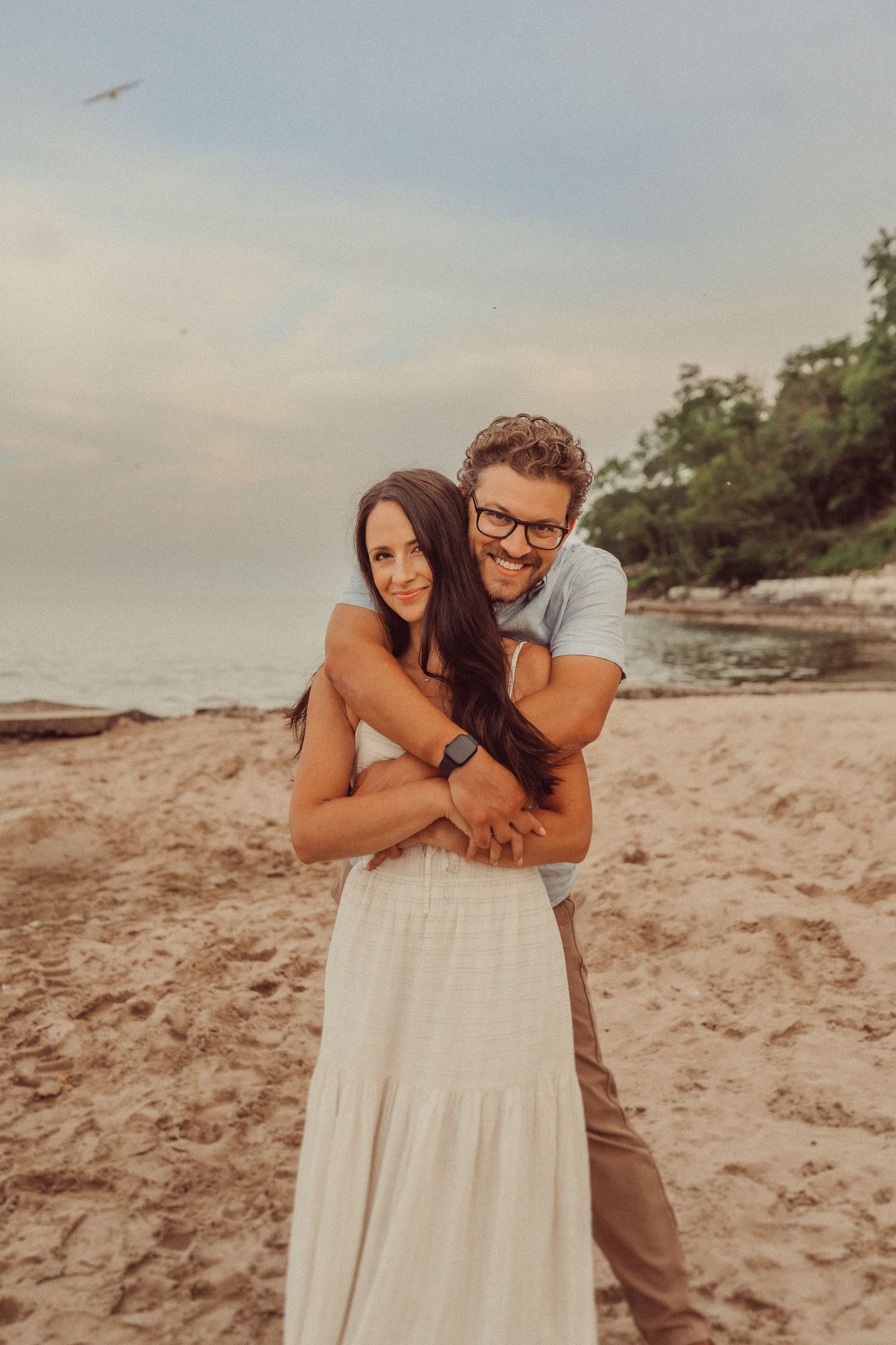 Loving embrace on beach with warm sunset lighting and coastal cliffs in background.