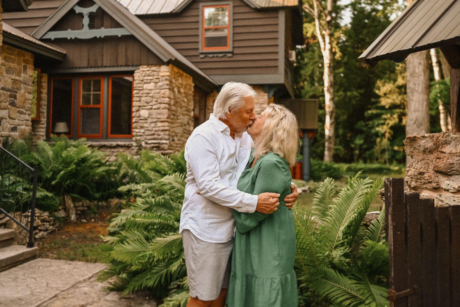 Senior couple share intimate moment outdoors by stone and wood cottage surrounded by ferns.