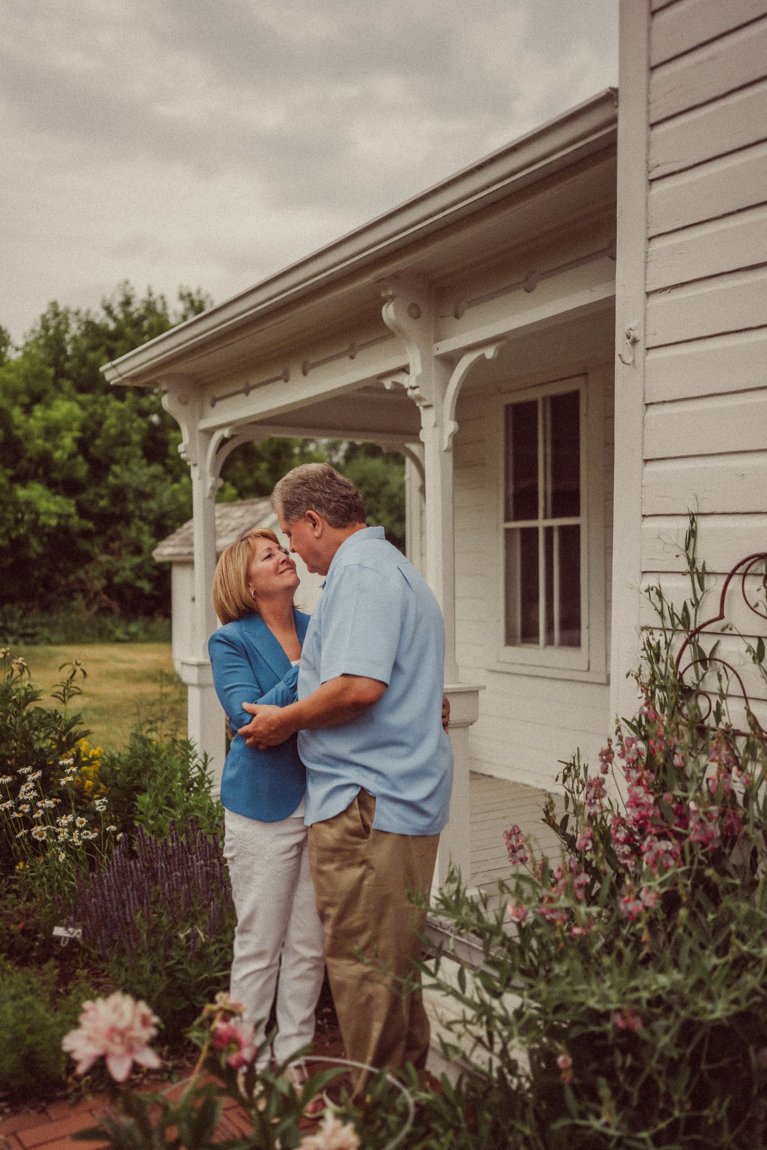 A loving couple shares a tender moment in front of a white farmhouse with ornate porch details and flowering garden.