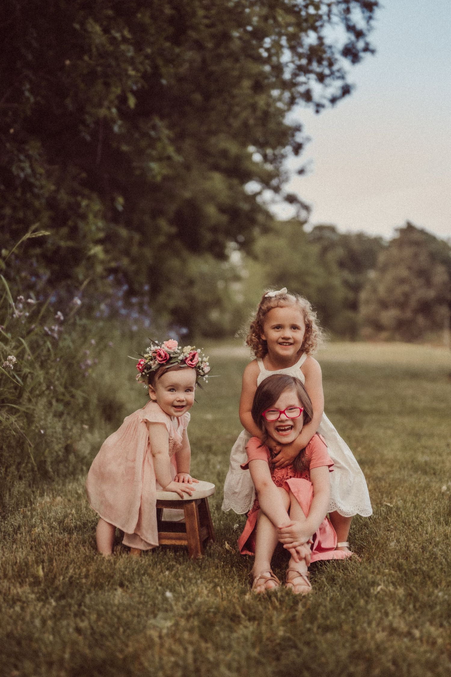 Three children in pink and white dresses pose together outdoors with a small wooden stool.