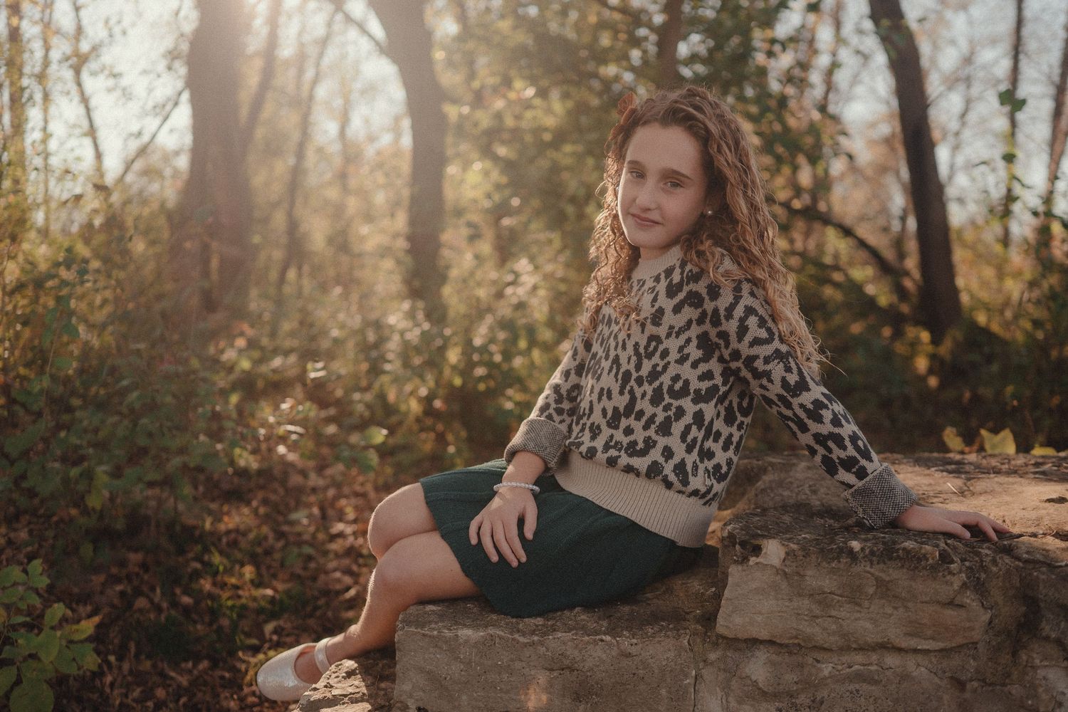 Person in leopard print sweater sits on rocks in sunlit autumn forest during golden hour.