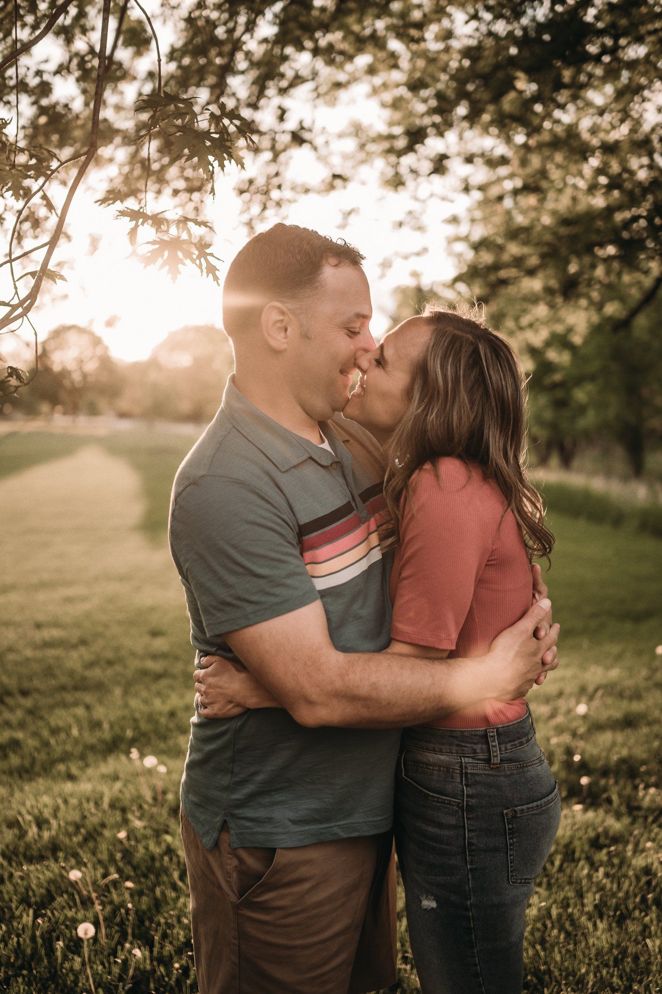 Intimate couple sharing a tender kiss in a park during sunset with trees in the background.