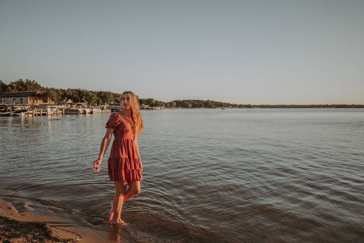 Woman in coral dress stands at the edge of a lake during sunset.