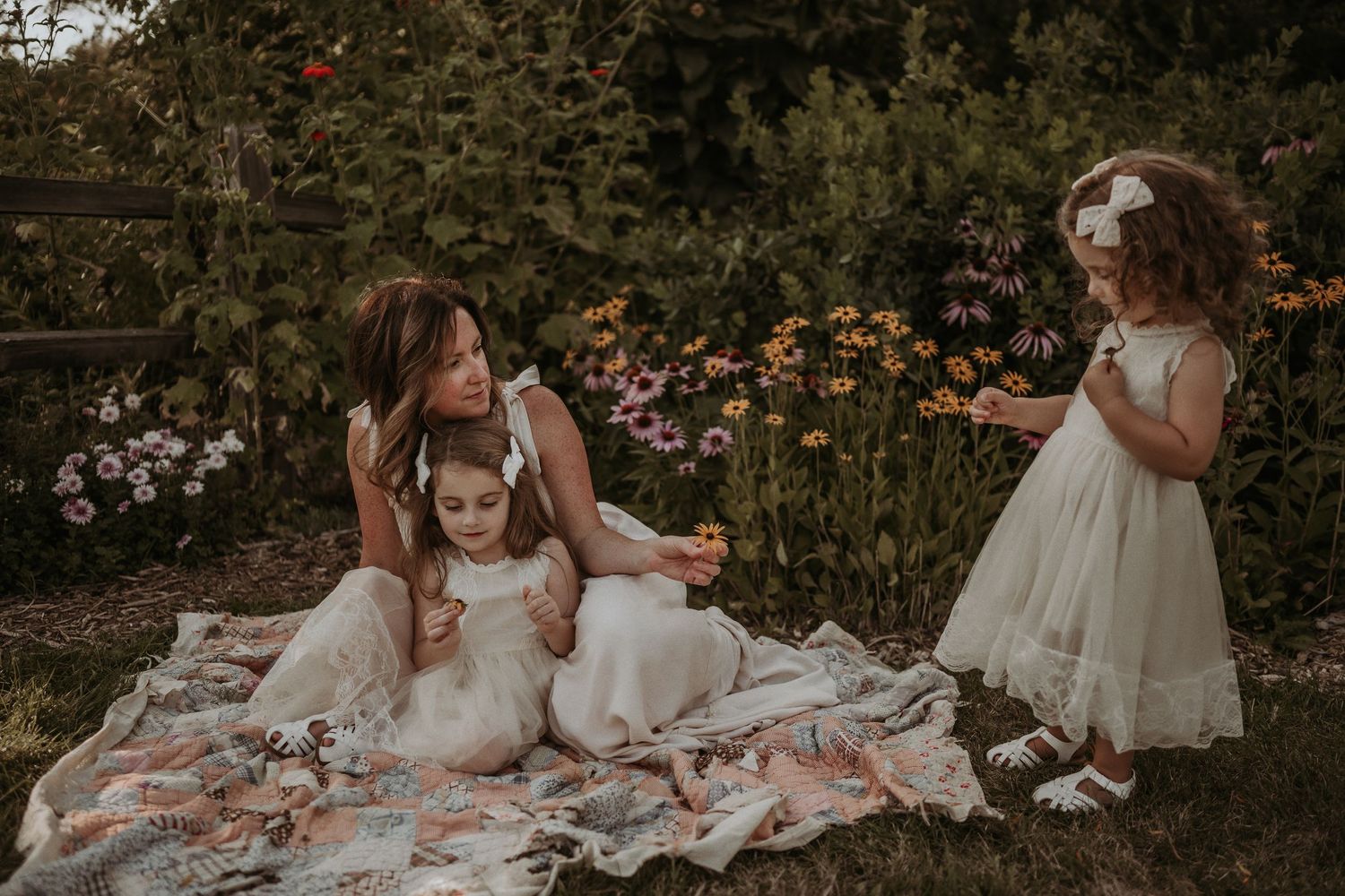 A garden photoshoot features people in white dresses sitting on a floral blanket.