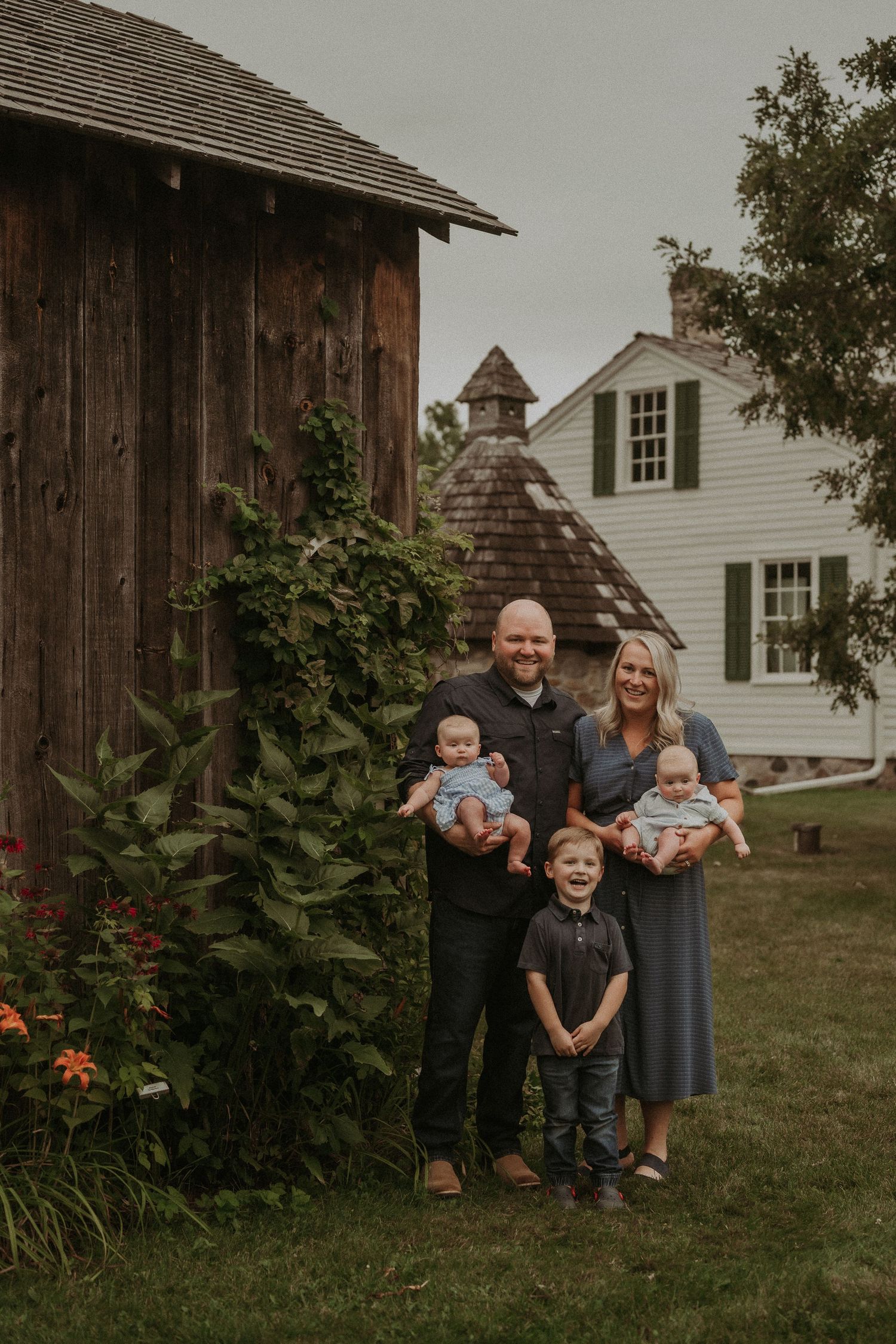 Small family poses for portrait near rustic barn and white farmhouse on cloudy day.