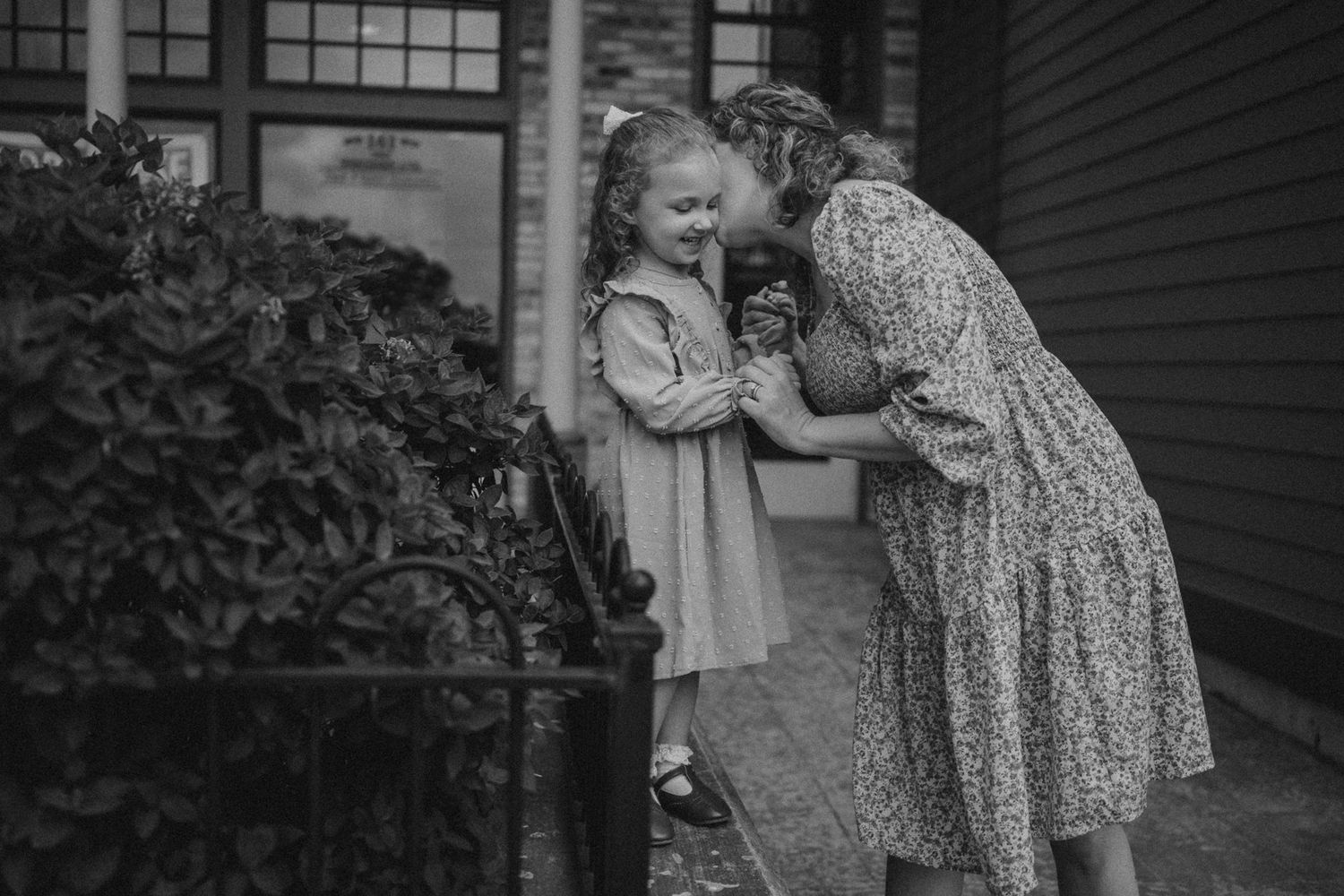 A black and white photo of a tender moment between two people next to a house with bushes.