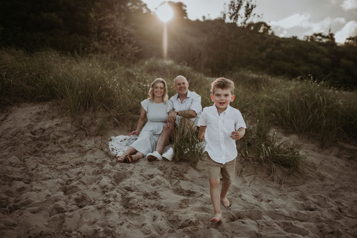 Child runs playfully on beach dunes while family watches in background during sunset.
