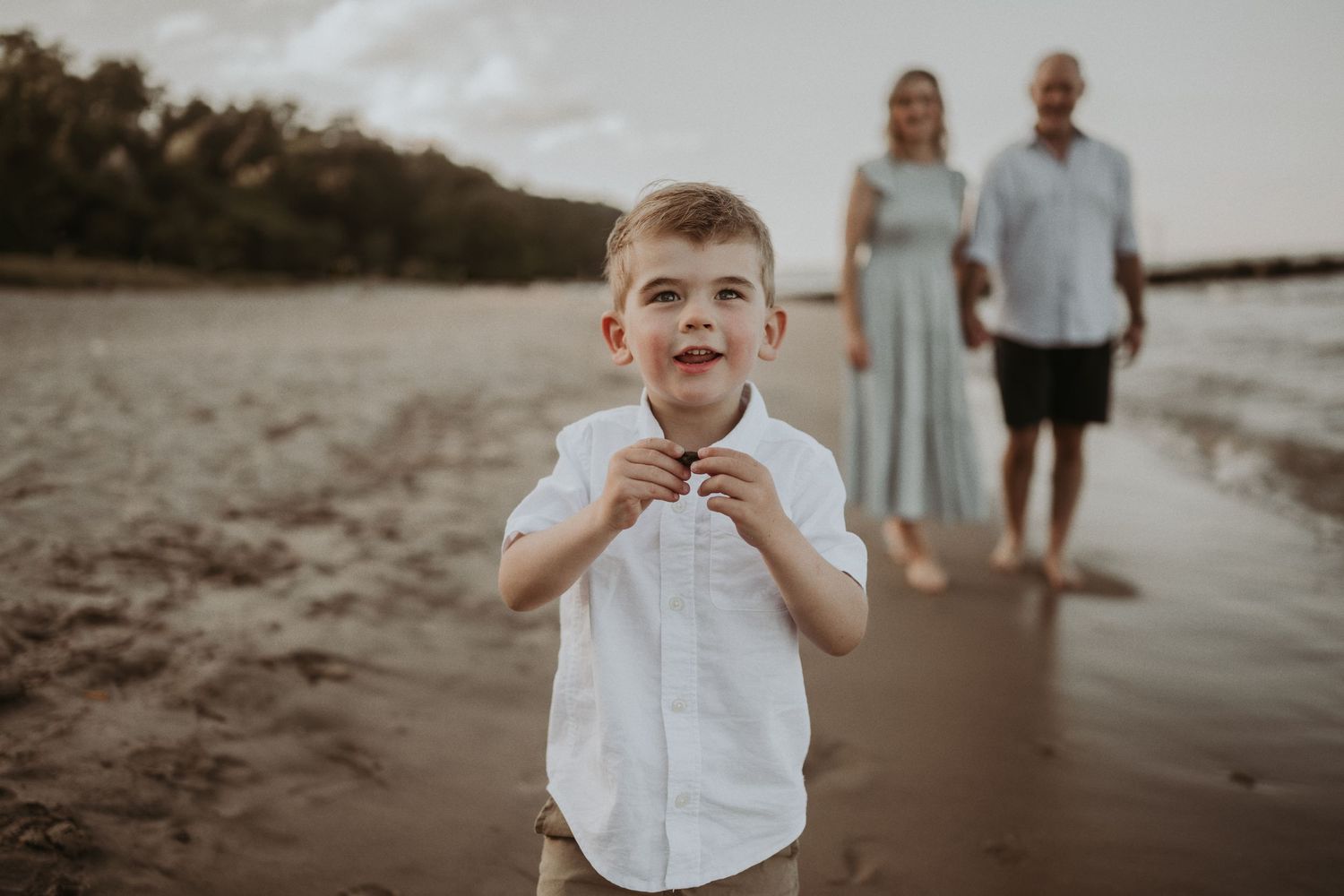 A young child in a white shirt poses on a beach at sunset while two people stand in the background.