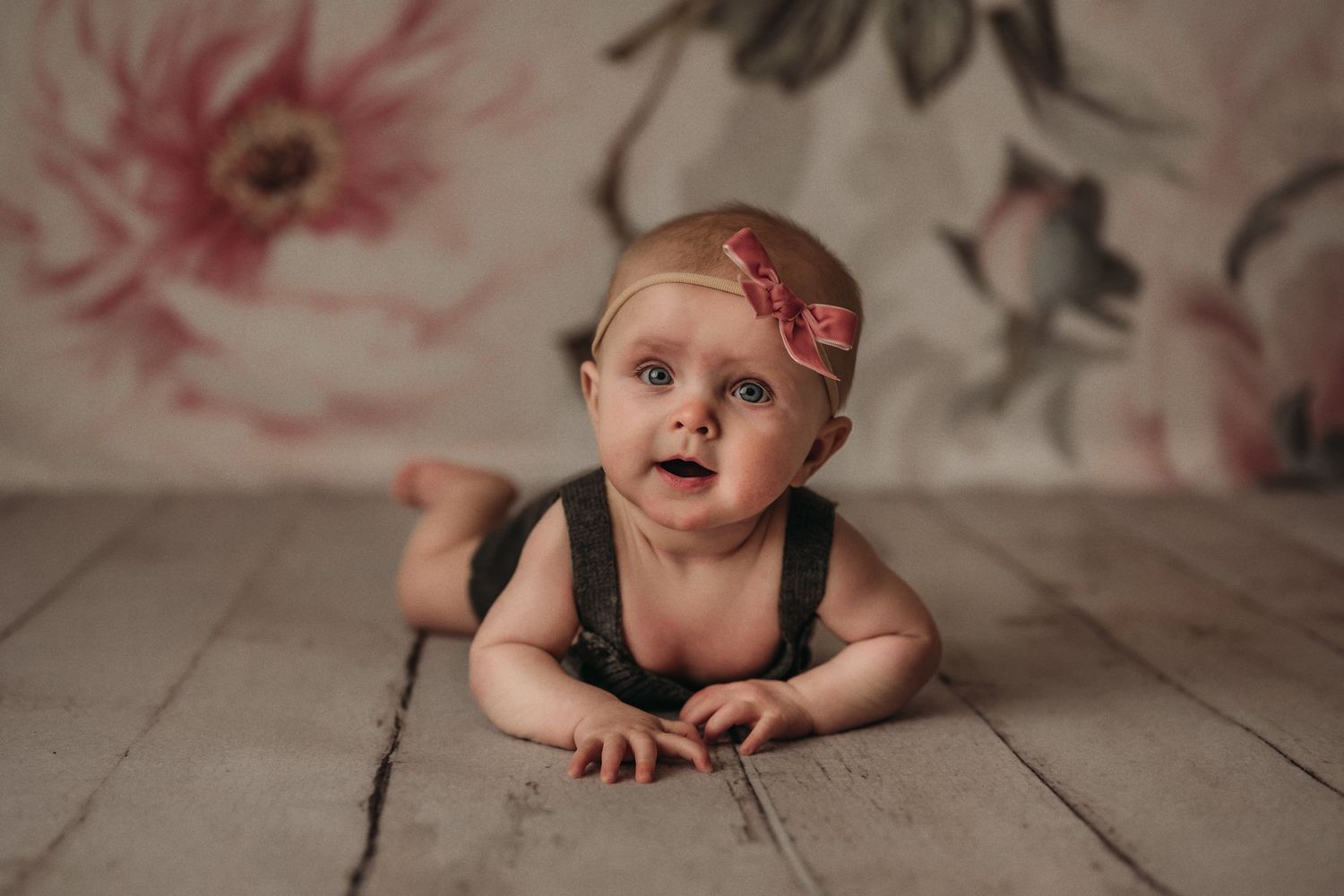 Baby in dark romper and headband lying on wooden floor with floral backdrop.