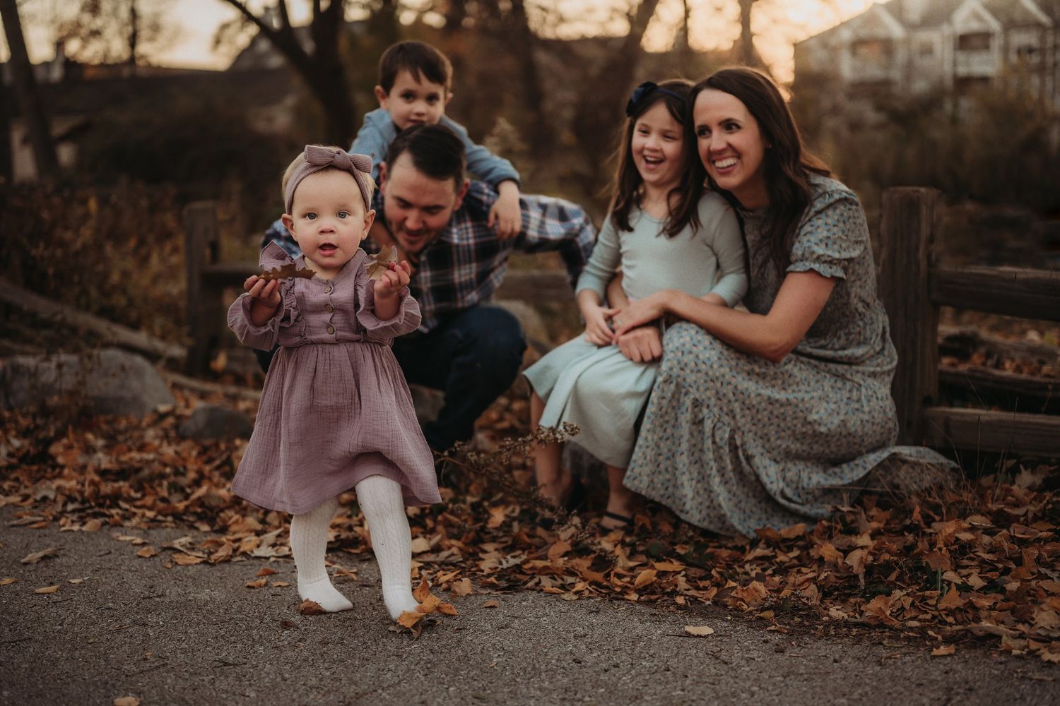 A toddler dances in a dress while others watch from their seat on fallen leaves.