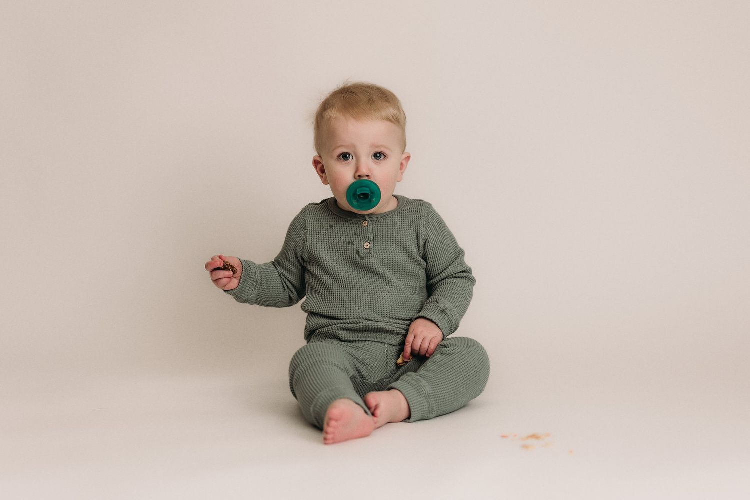 Toddler sitting in sage green ribbed outfit with pacifier against light background.