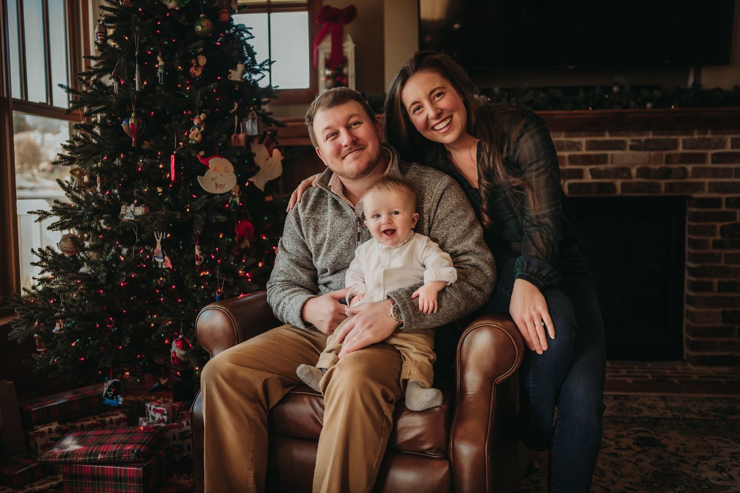 Family of three poses for a warm holiday portrait by their decorated Christmas tree and brick fireplace.