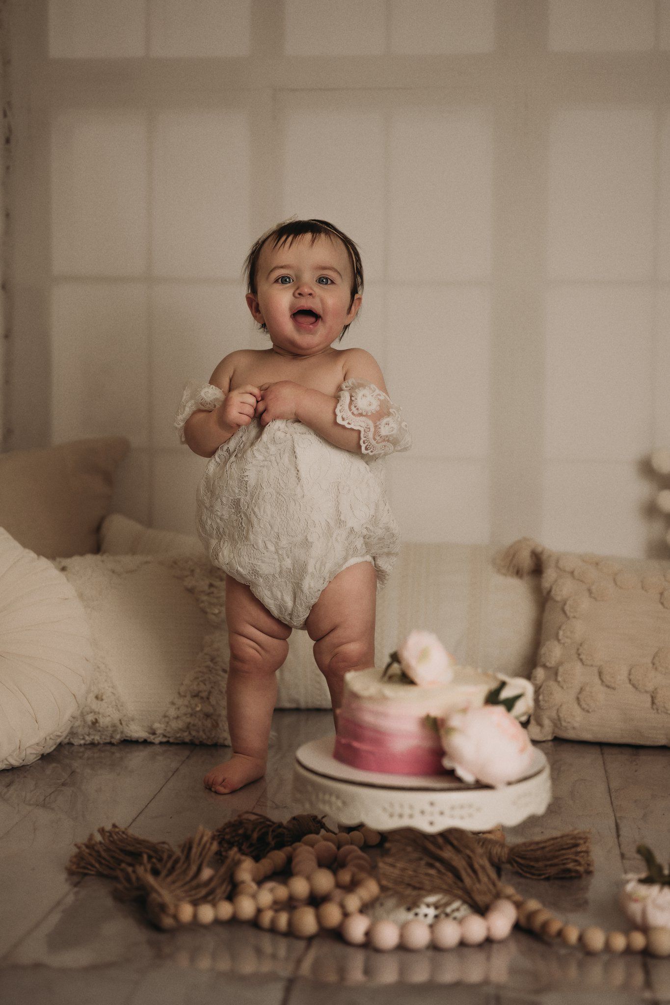 Baby in white lace outfit smiling beside pink cake and wooden bead decorations.