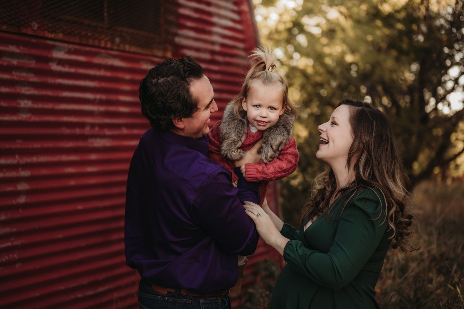 Family sharing playful moment together by red barn wall in warm sunlight.