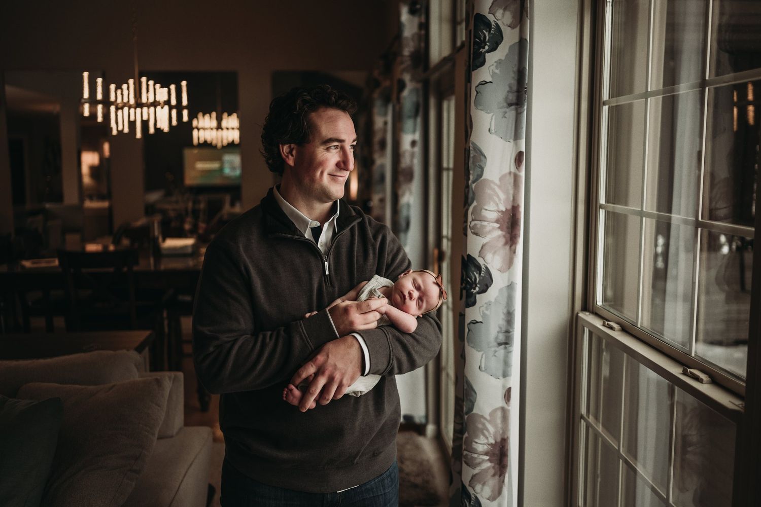 Parent wearing dark jacket smiles warmly while cradling newborn by sunny window.