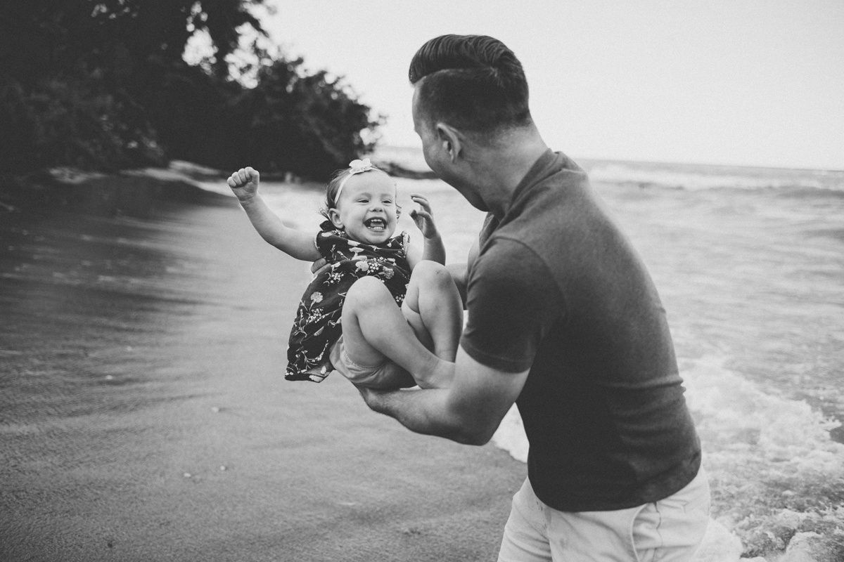 A playful black and white capture of a parent lifting a laughing child by the beach.