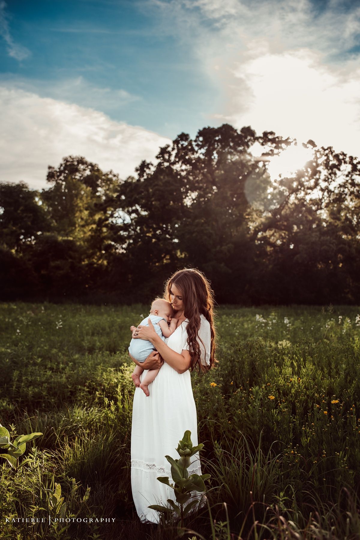 Mother holding baby in white dress against sunset in meadow with trees.