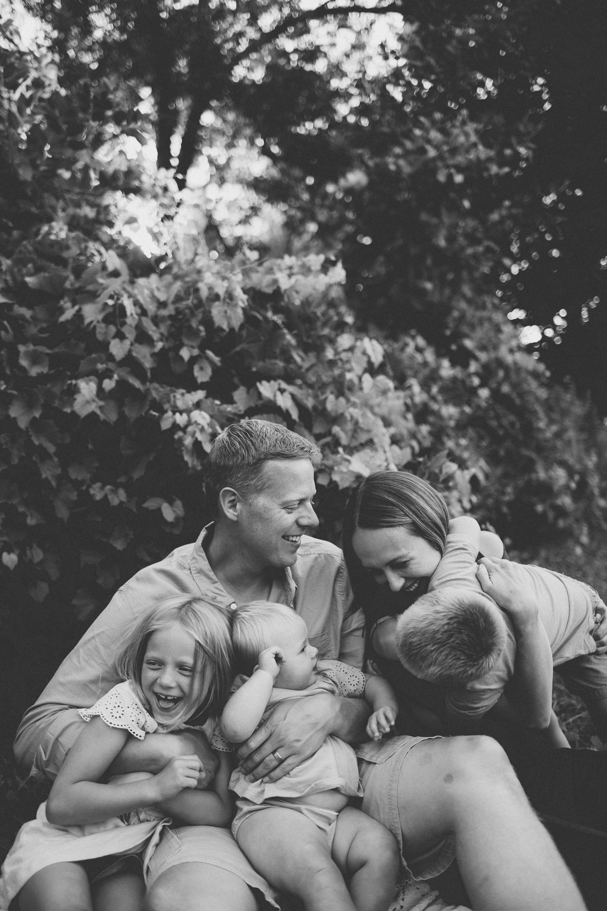 Black and white portrait of family sharing laughter and cuddles on blanket outdoors.