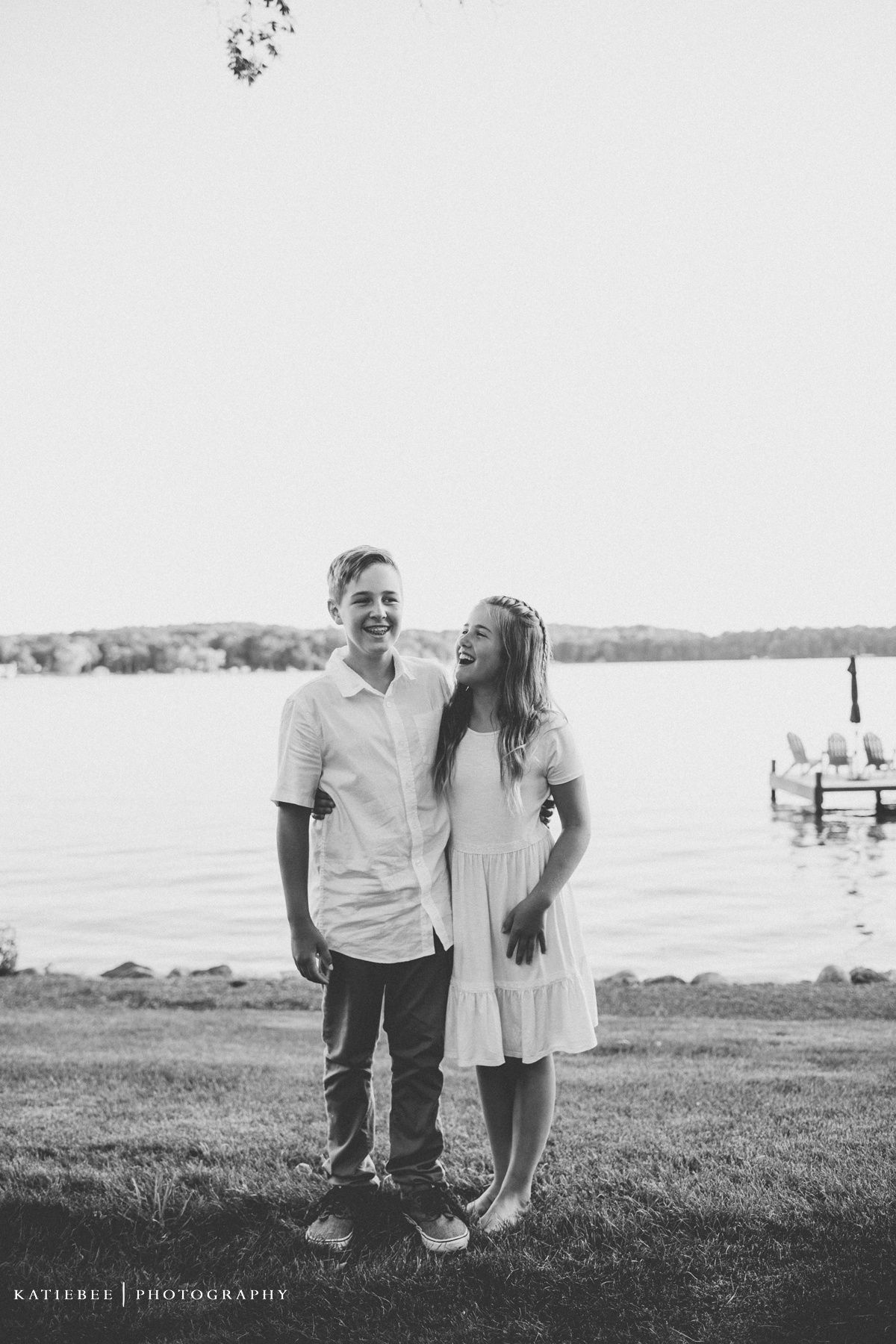 Black and white portrait of two people standing by lake with dock in background.
