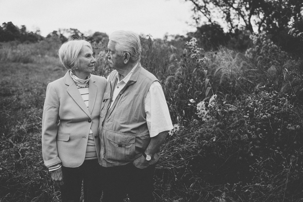 A black and white portrait of a couple standing together outdoors among bushes.