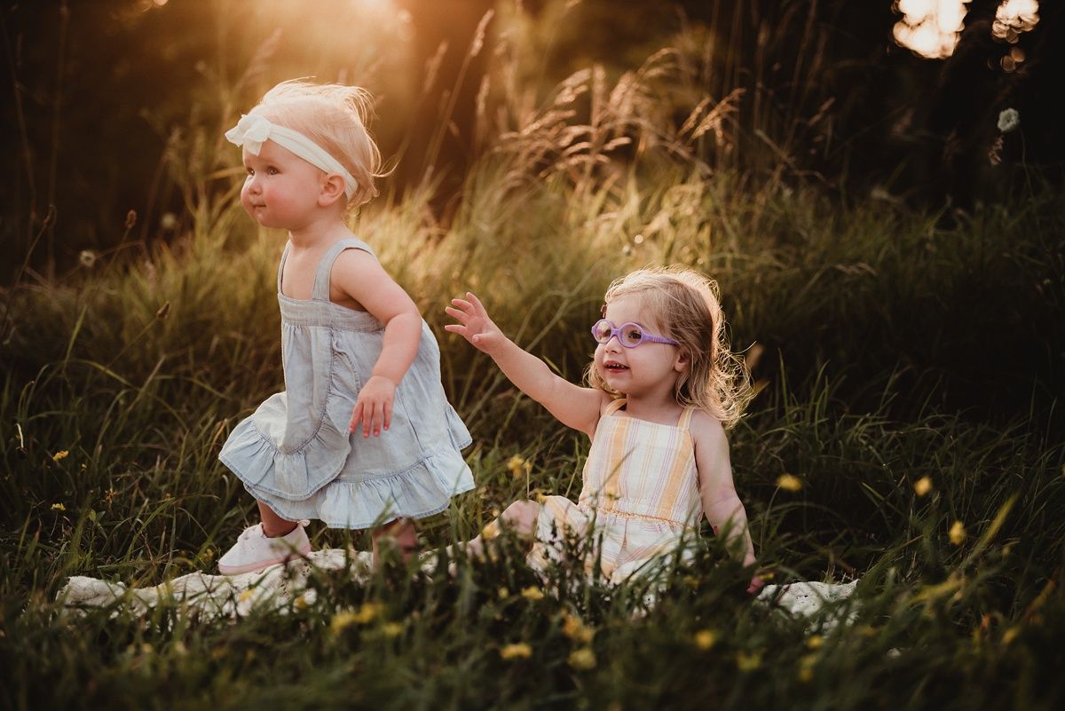 Two children in light dresses play together in a field at sunset.